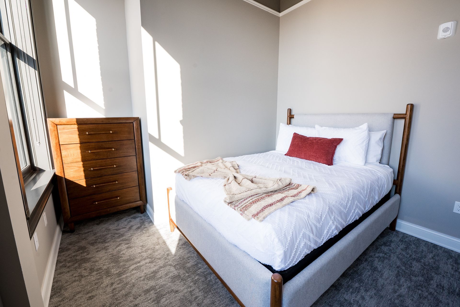 Bright bedroom with a white bed, red pillow, gray carpet, and a wooden dresser by the window.