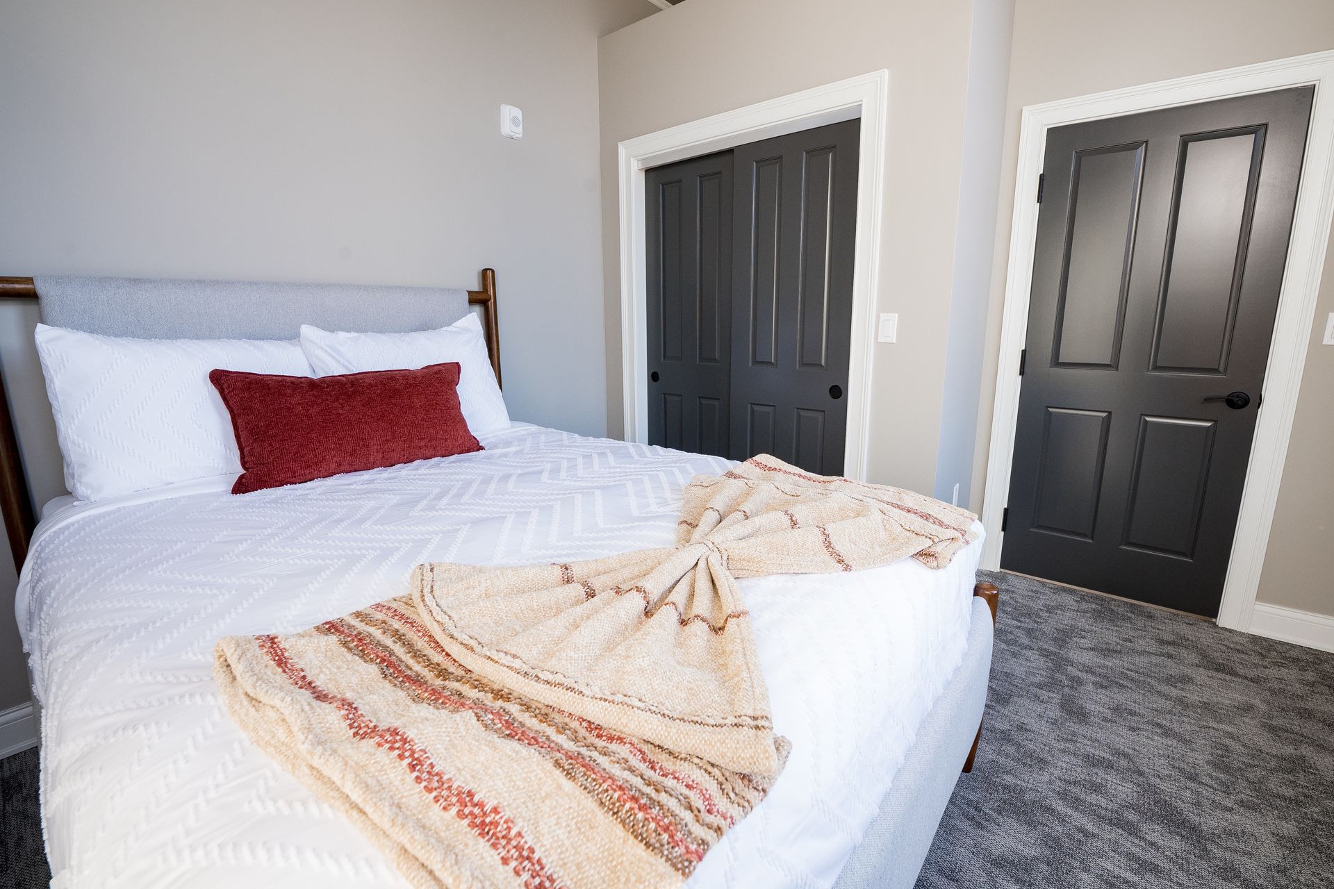 Bright bedroom with a made white bed, red pillow, and gray carpet beside dark closet doors.