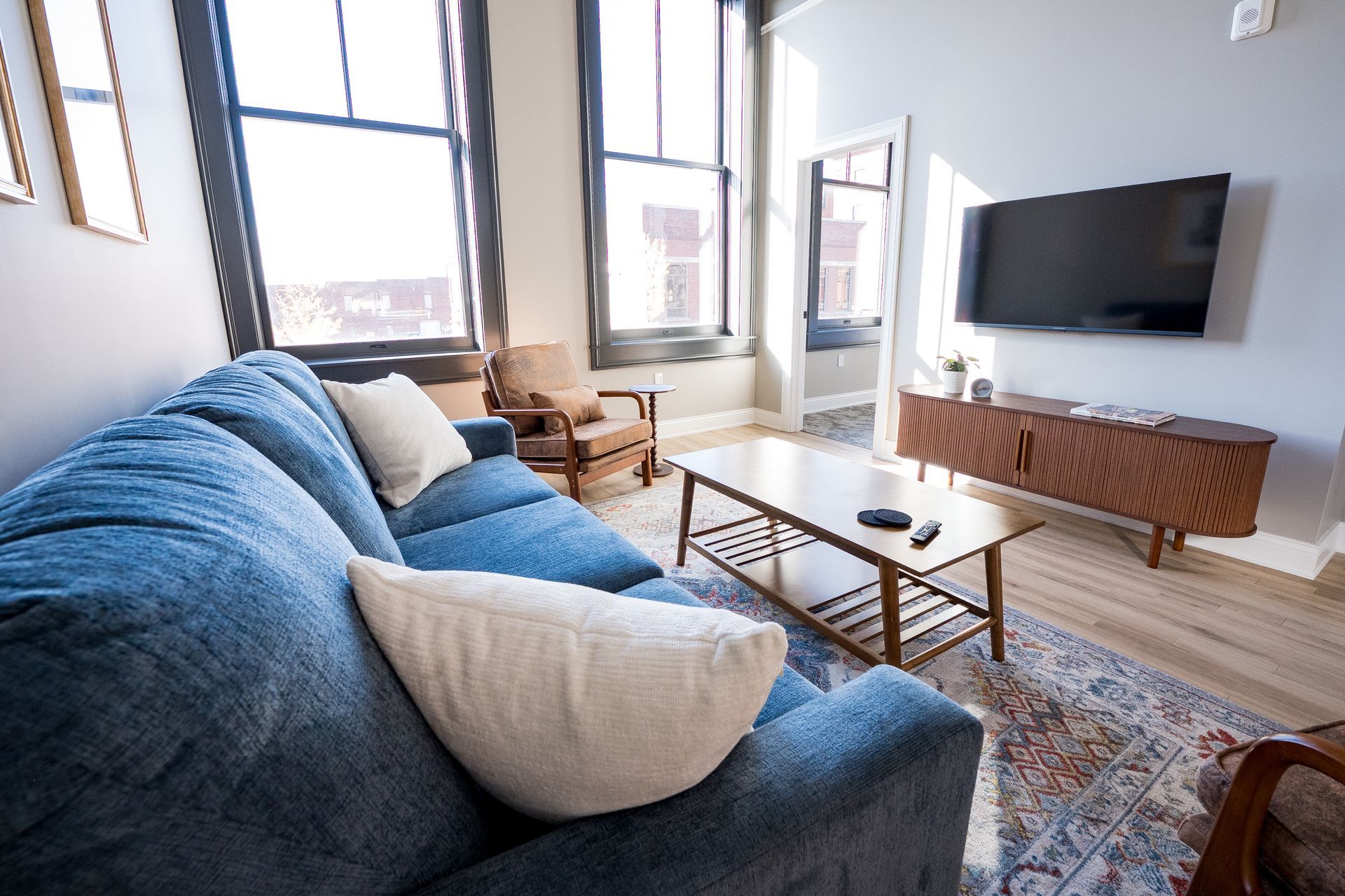 Bright living room with blue sofa, white armchair, wall-mounted TV, and large windows