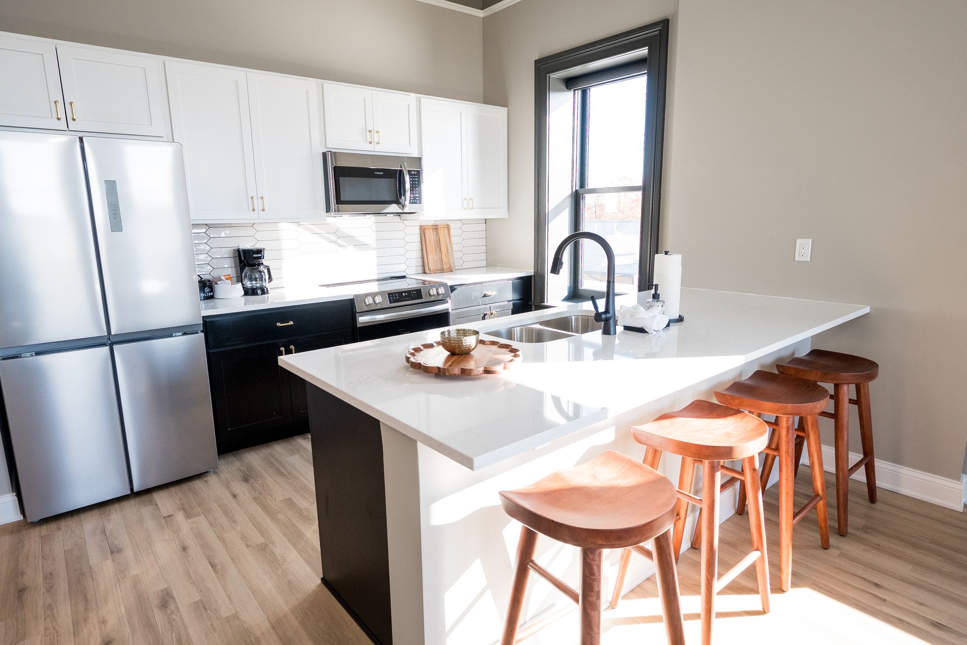 Modern kitchen with stainless appliances, white cabinets, island sink, and wooden stools by a bright window