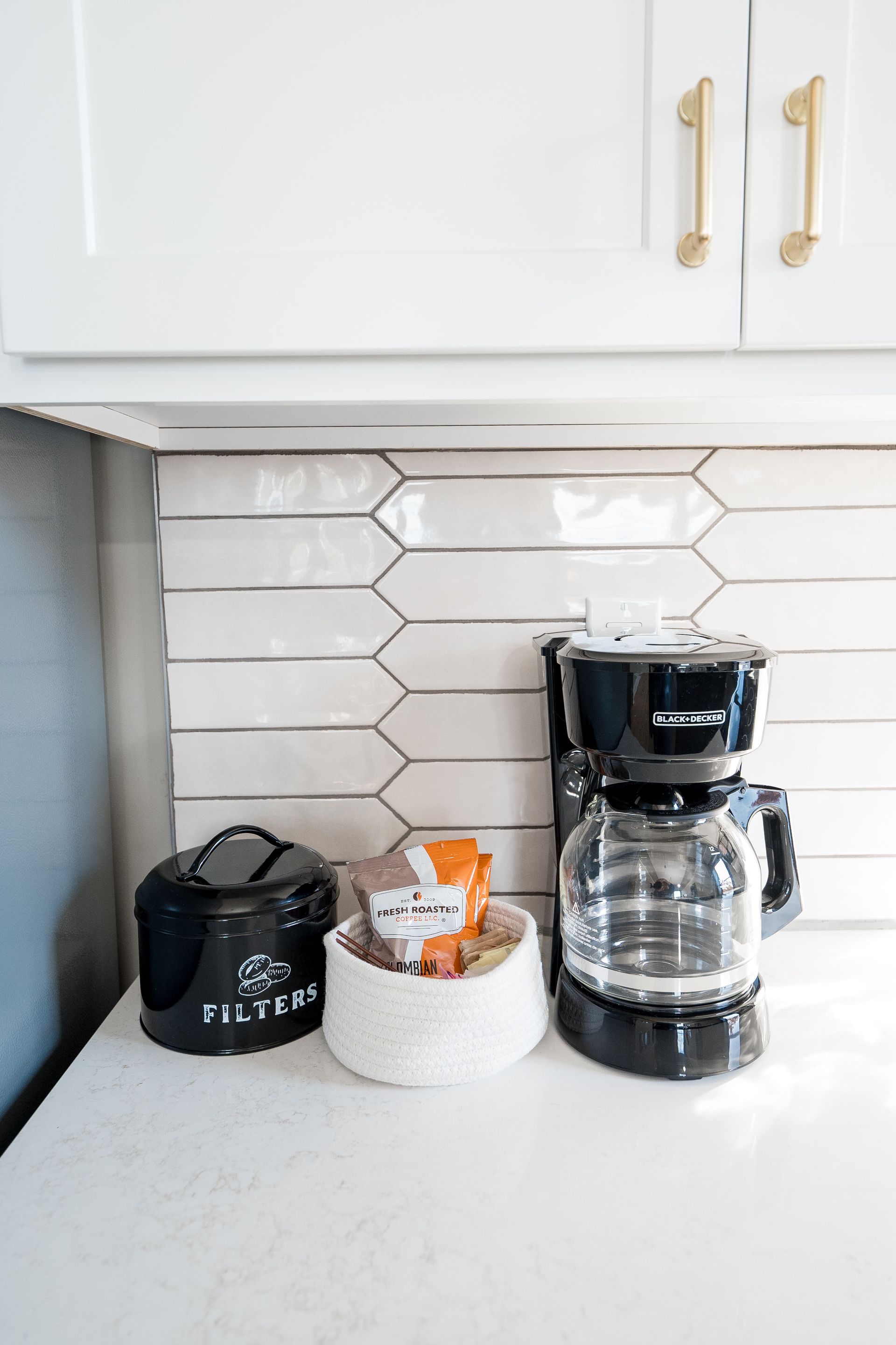 Kitchen counter with black coffee maker, small black appliance, and white basket under white cabinets and tile backsplash