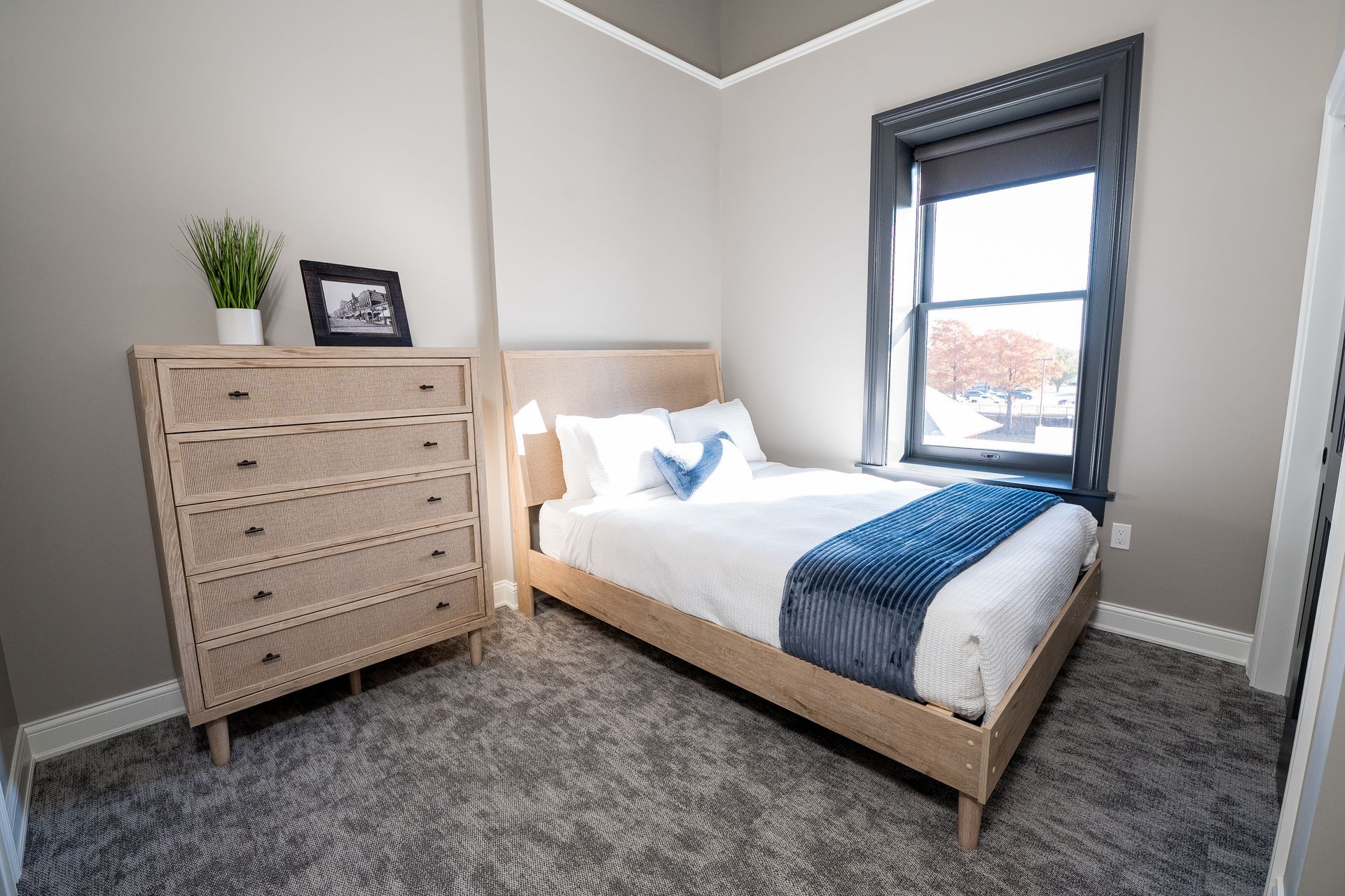 Small bedroom with light wood bed, dresser, gray carpet, and a window with dark curtain.