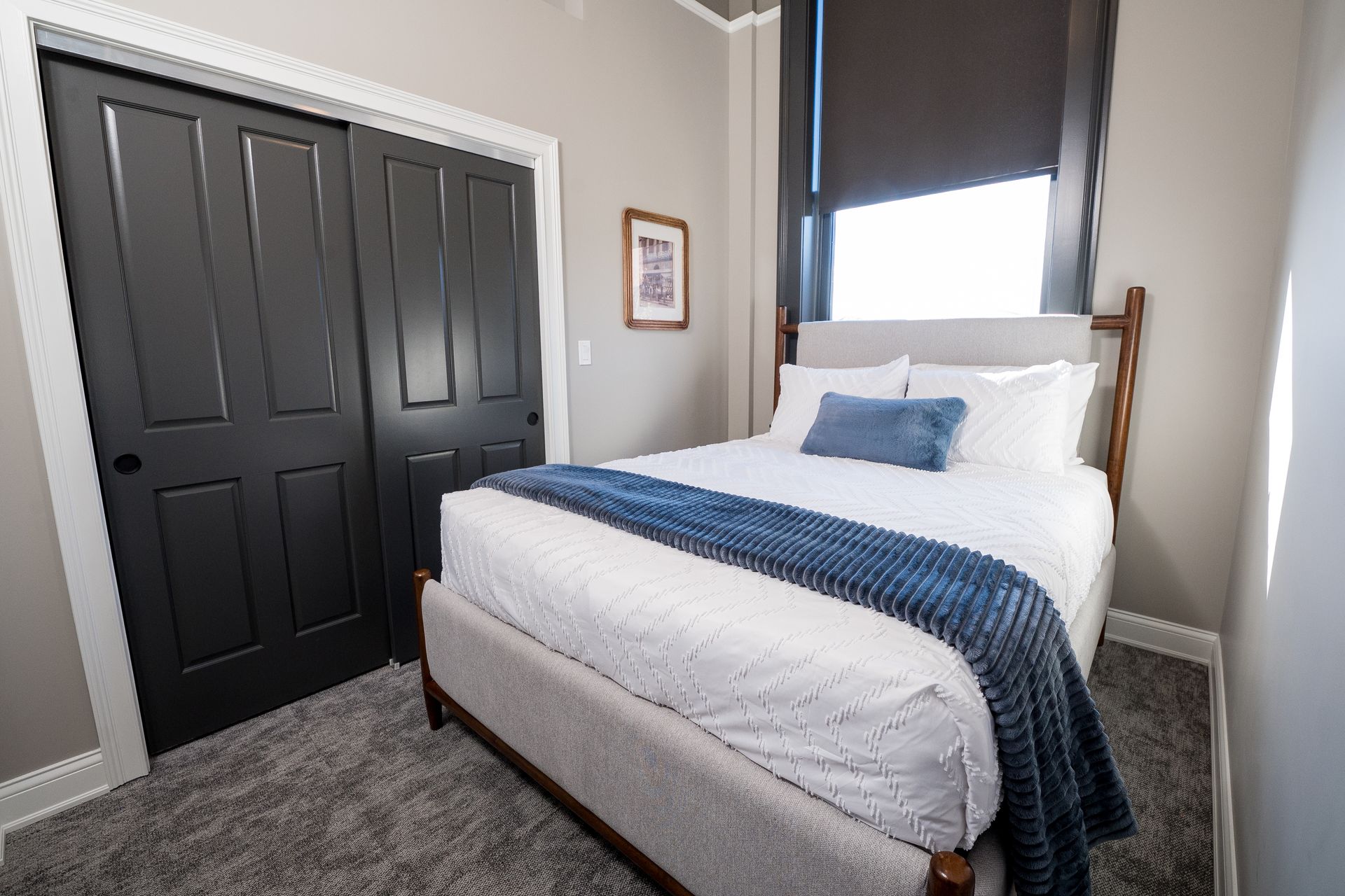 Small bedroom with a white bed, navy throw, gray carpet, and black closet doors.