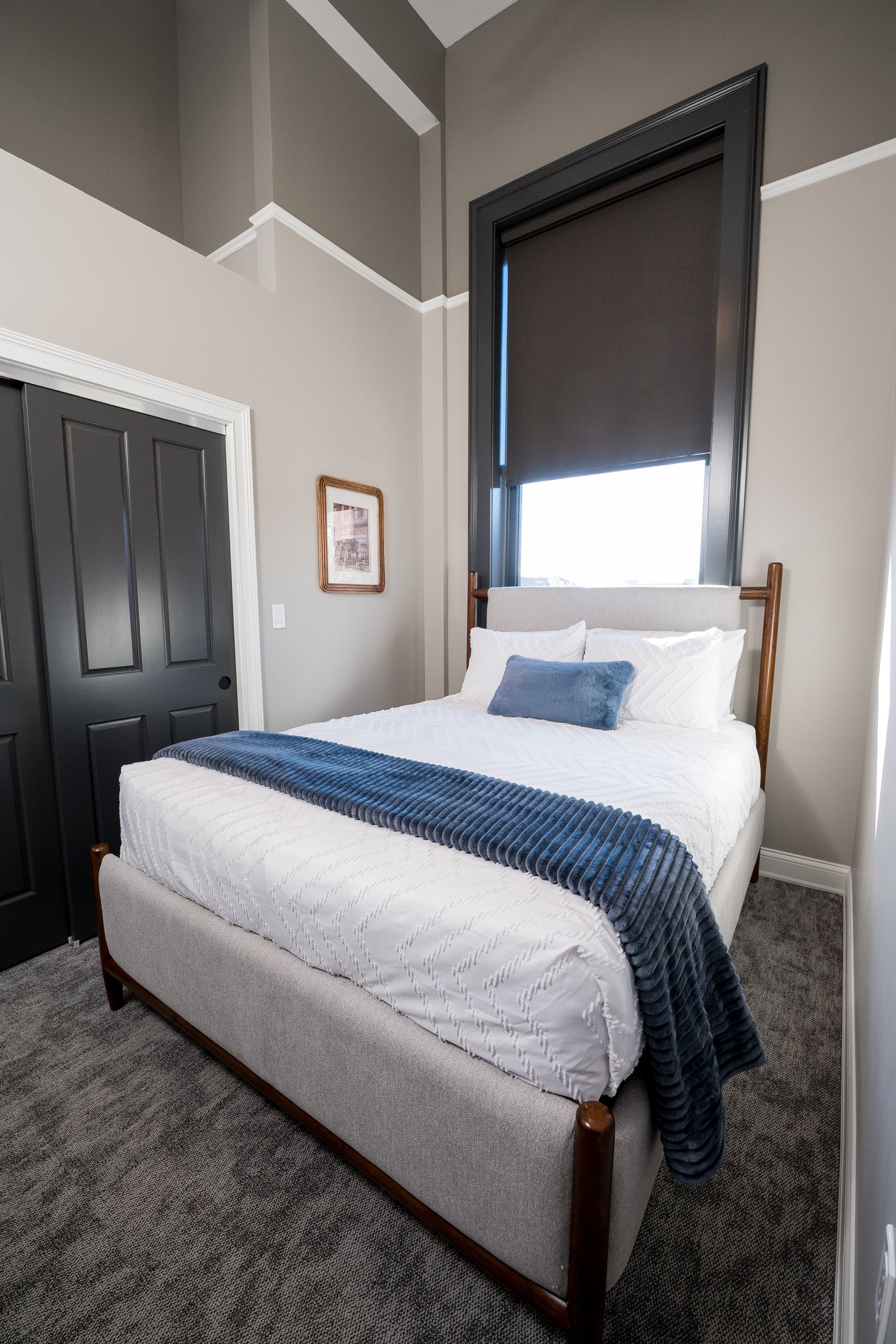 Small bedroom with a neatly made white bed, blue throw, dark closet doors, and a window with dark blind.