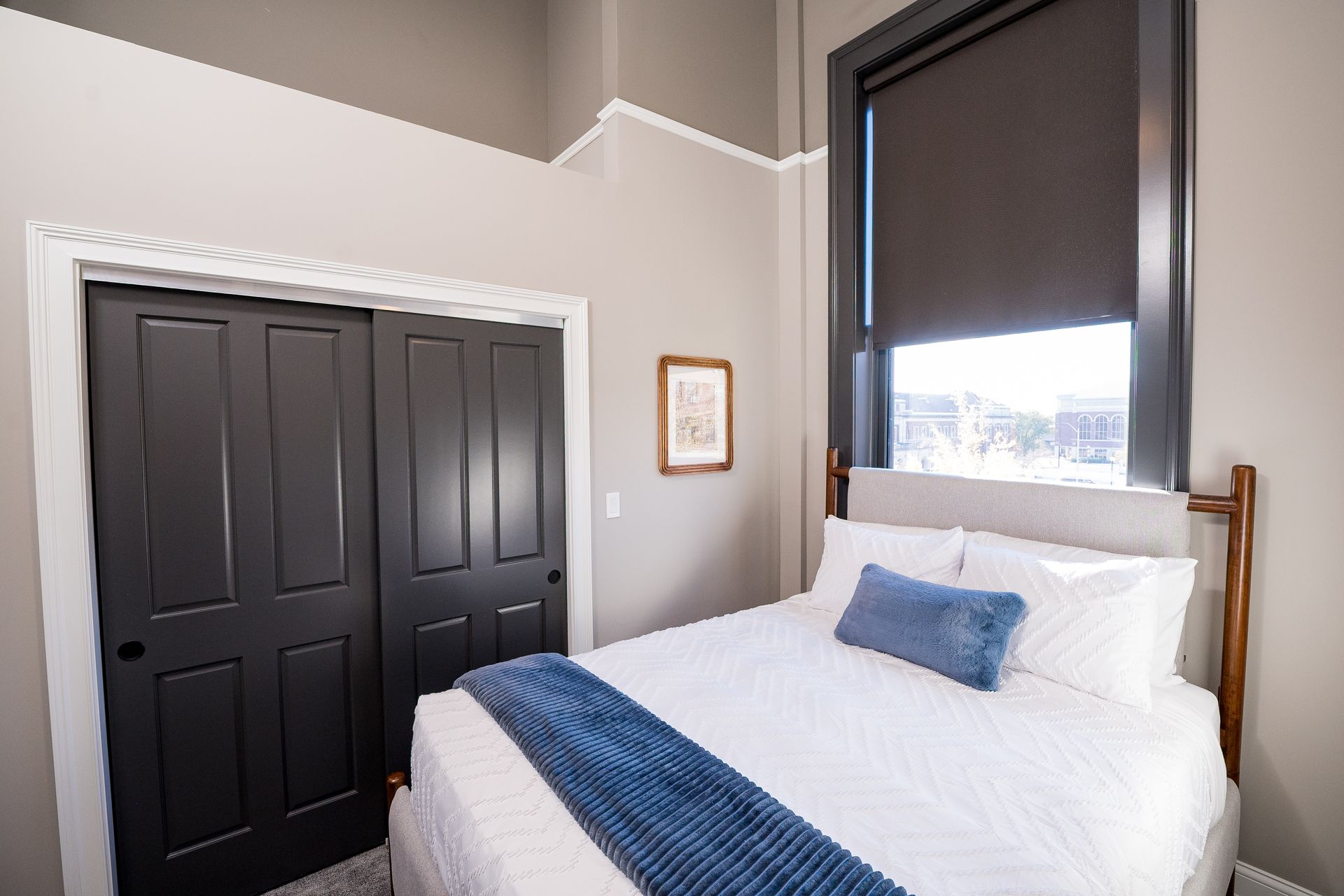 Small bedroom with a twin bed, blue accents, black closet doors, and a window with a dark shade.
