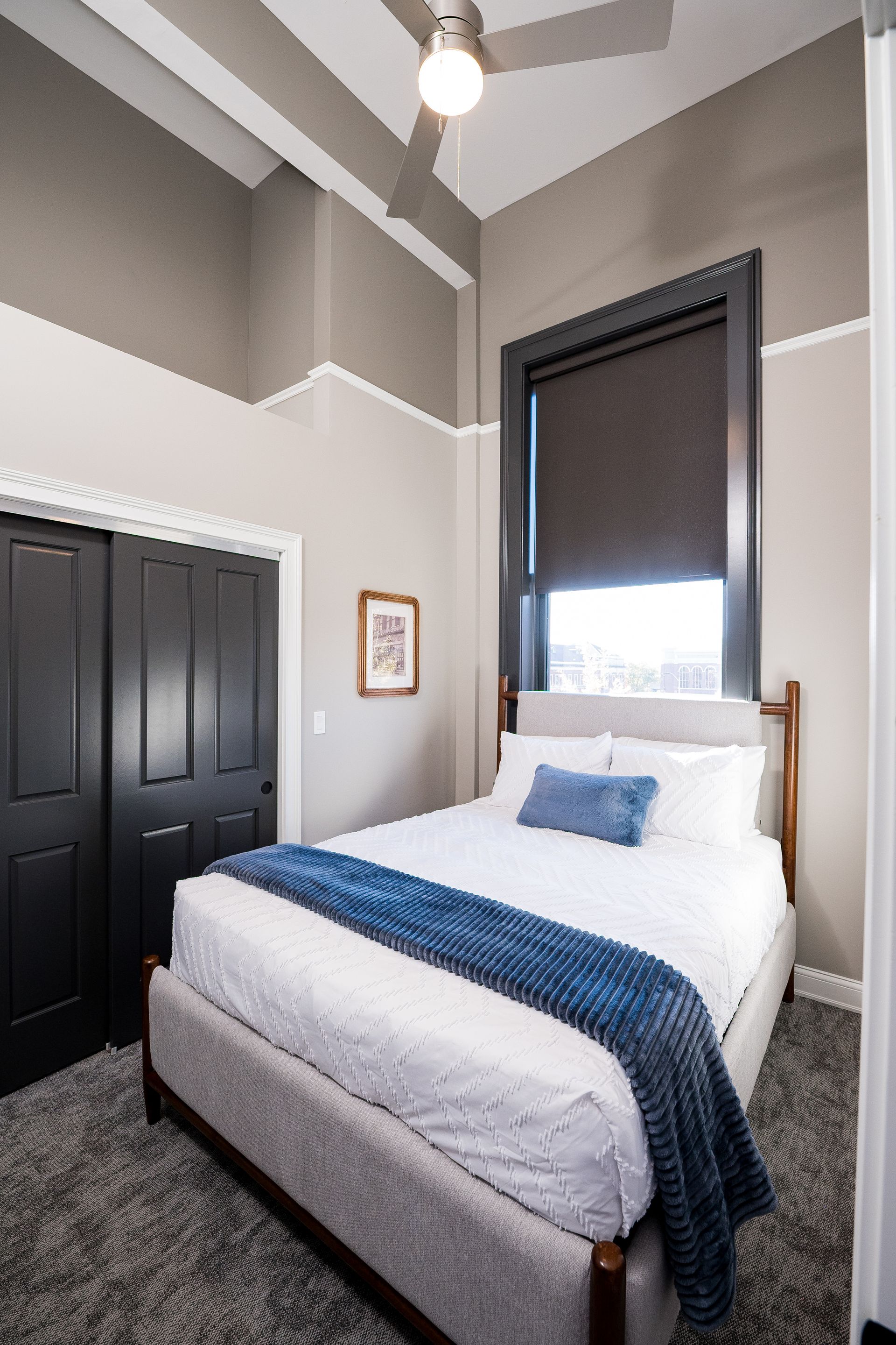 Small bedroom with a white bed, blue throw, black closet doors, and a tall window with a dark shade.