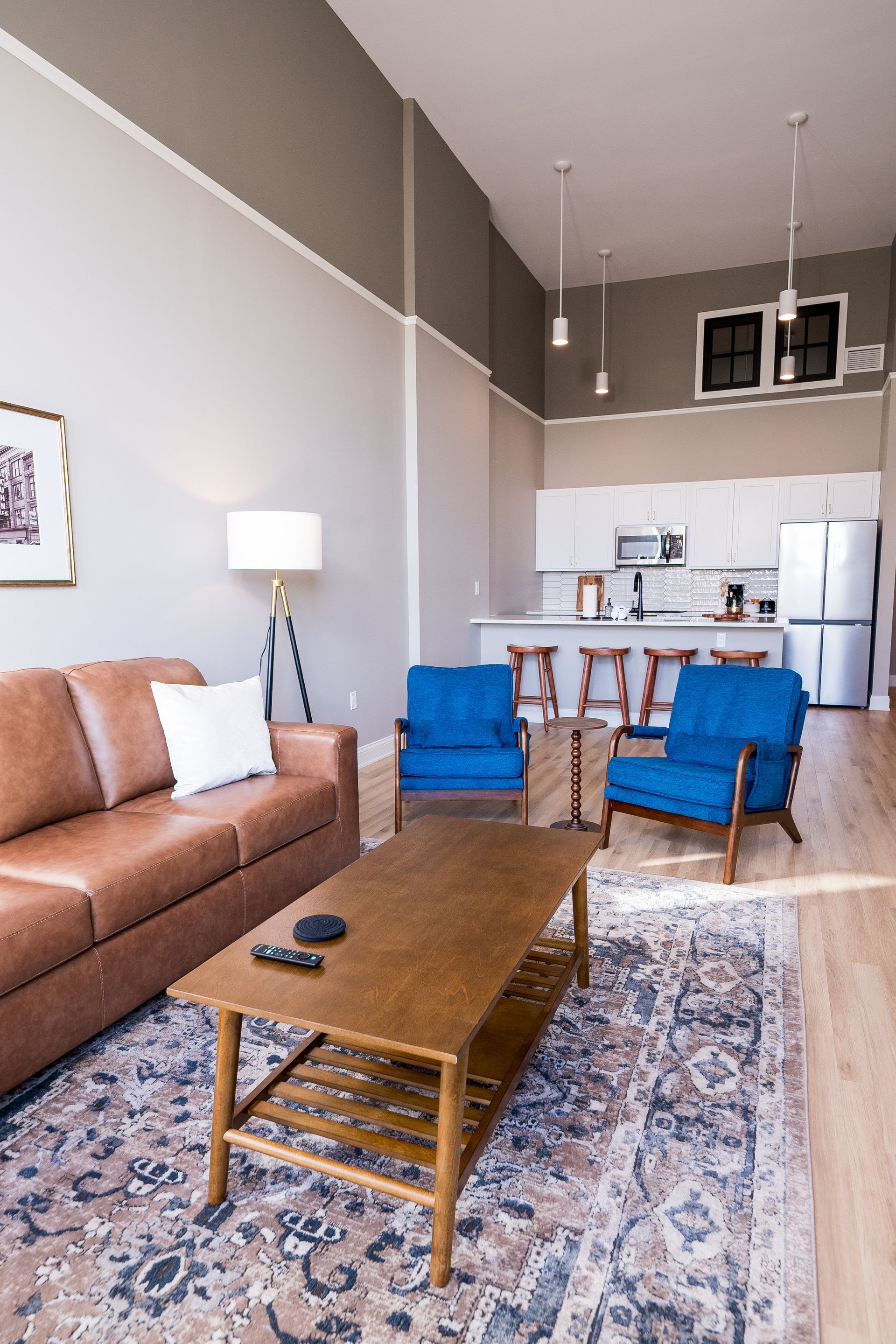 Bright living room with brown sofa, blue chairs, coffee table, patterned rug, and small kitchen area.