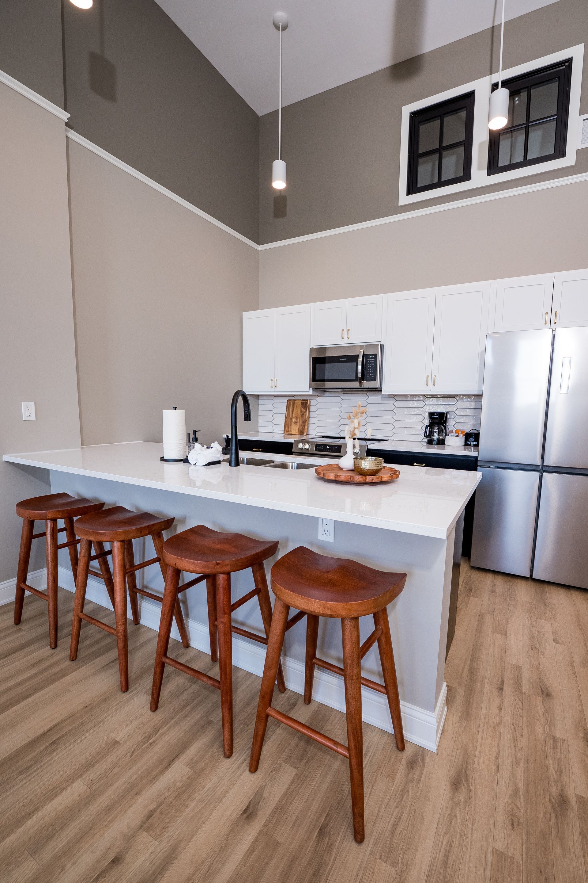 Modern kitchen with white island, wooden stools, stainless fridge, and high windows