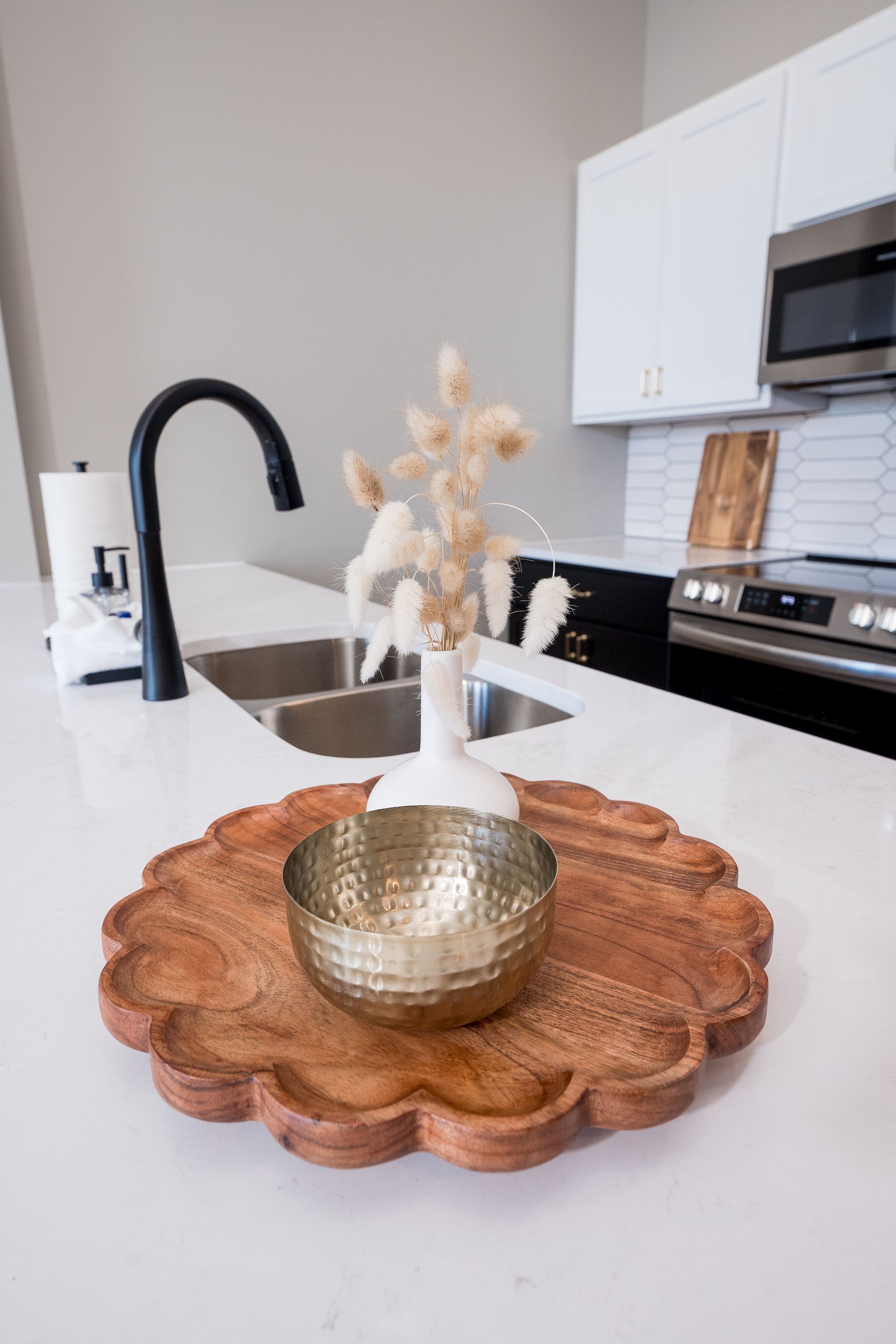 Kitchen island with a wooden tray, gold vase, and white flowers beside a black faucet