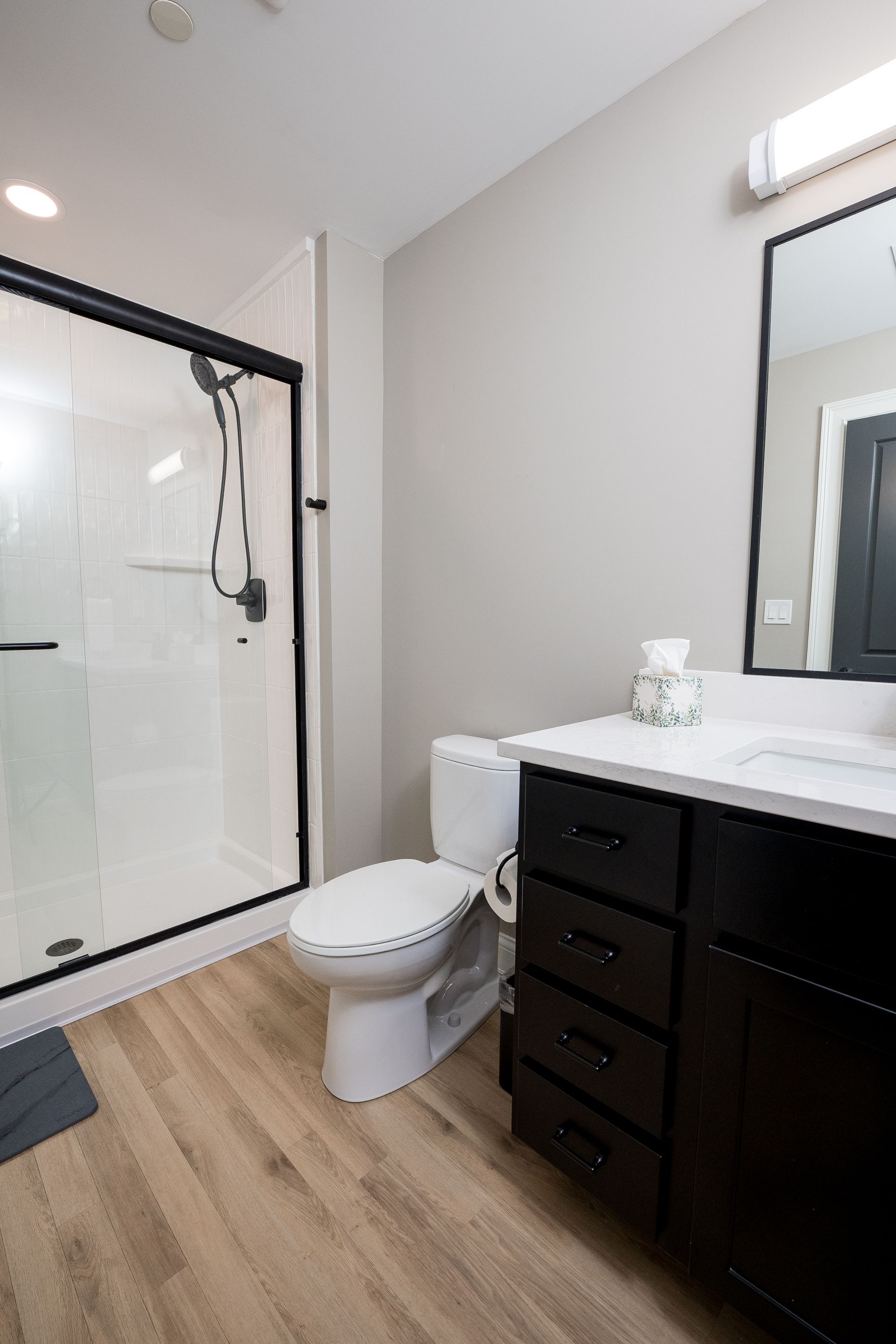 Modern bathroom with glass shower, toilet, dark vanity, and wood-look floor.