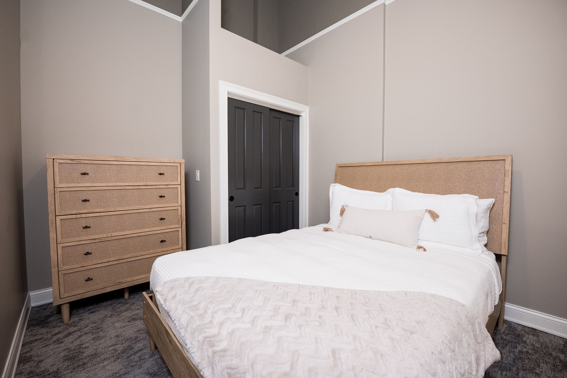 Minimal bedroom with white bed, woven dresser, and dark door against gray walls
