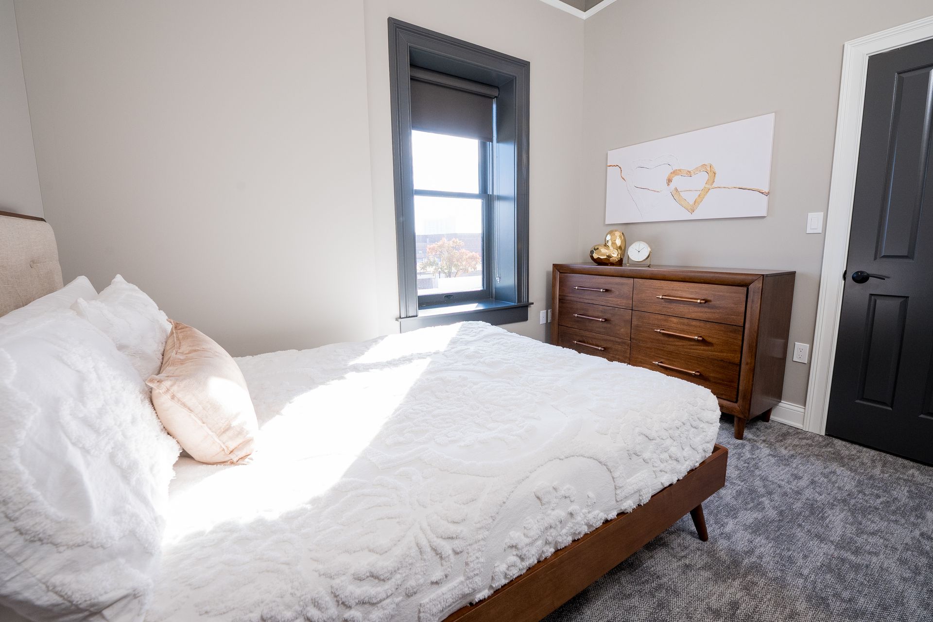 Bright bedroom with unmade white bed, dark window, wooden dresser, and black door.