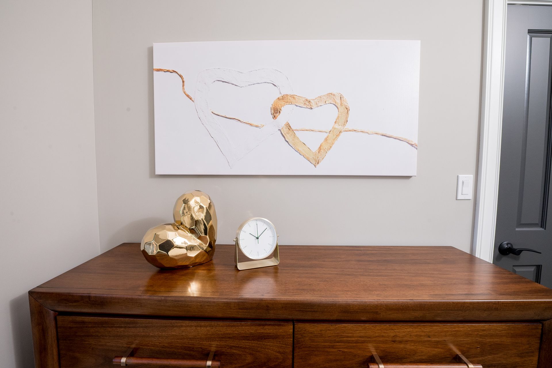 Wooden dresser with gold decor and a small clock beneath abstract heart wall art