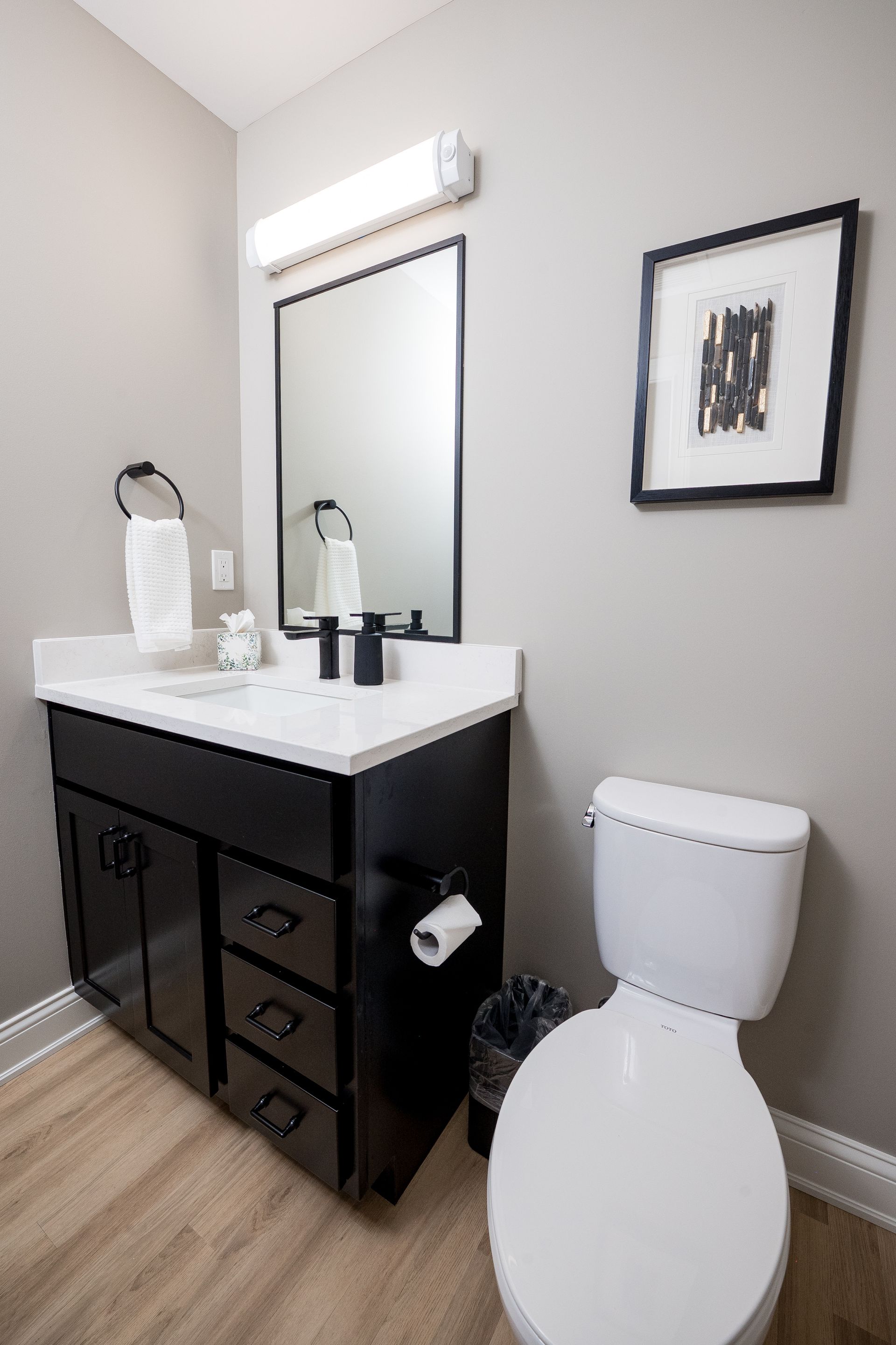 Bathroom with black vanity, white sink, mirror, and toilet against light gray walls.