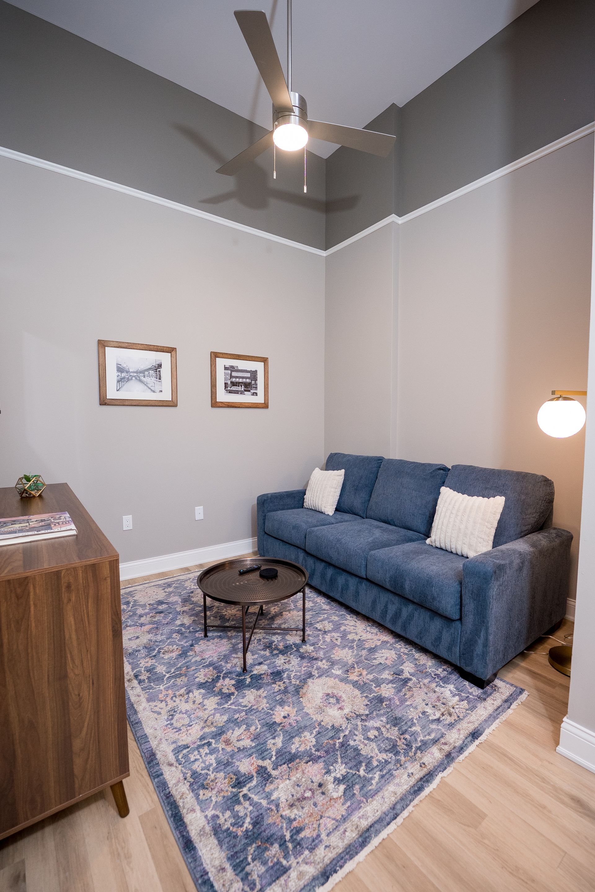 Small cozy living room with blue sofa, patterned rug, coffee table, floor lamp, and ceiling fan.