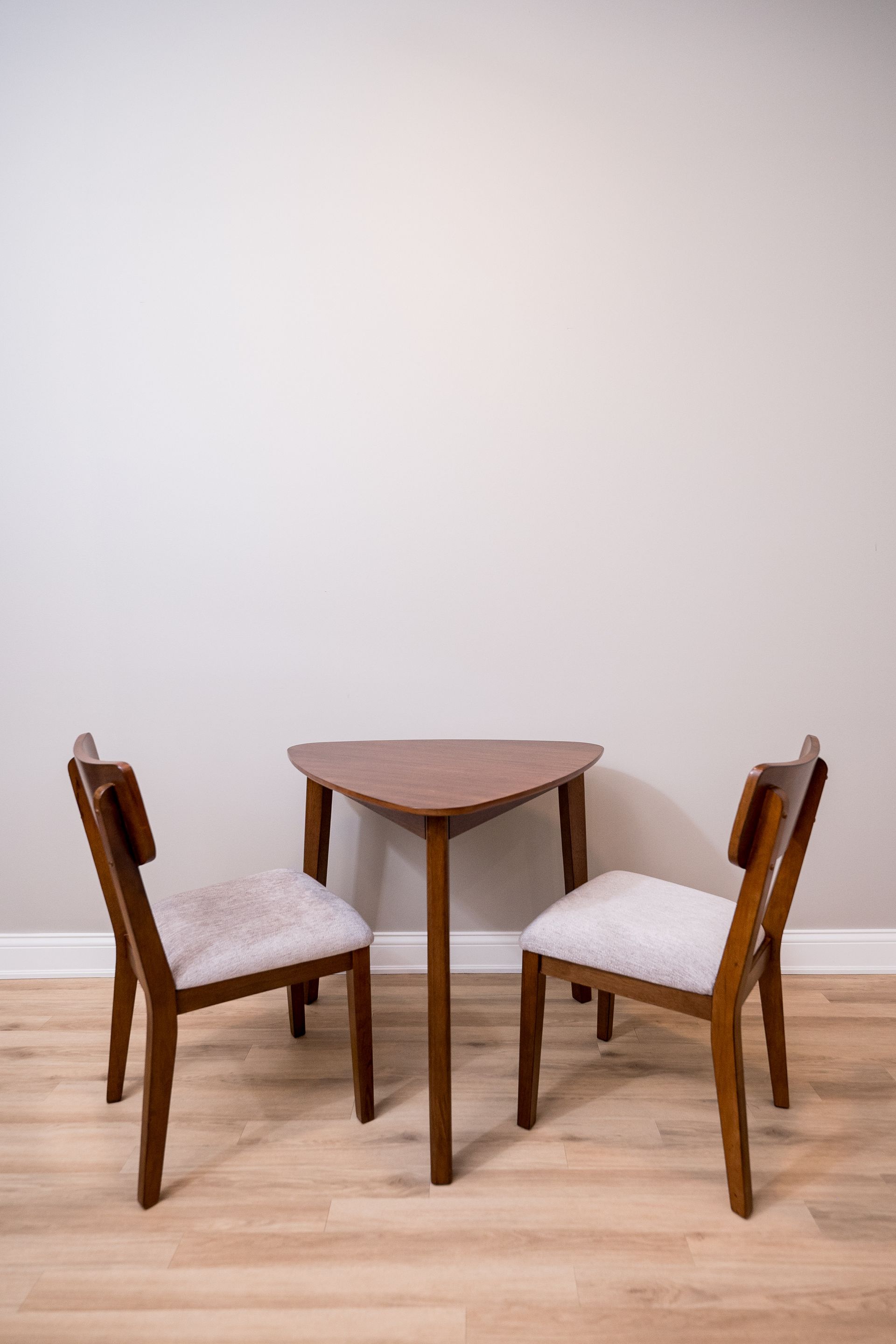 Small wooden table with two upholstered chairs on a light wood floor against a blank wall.