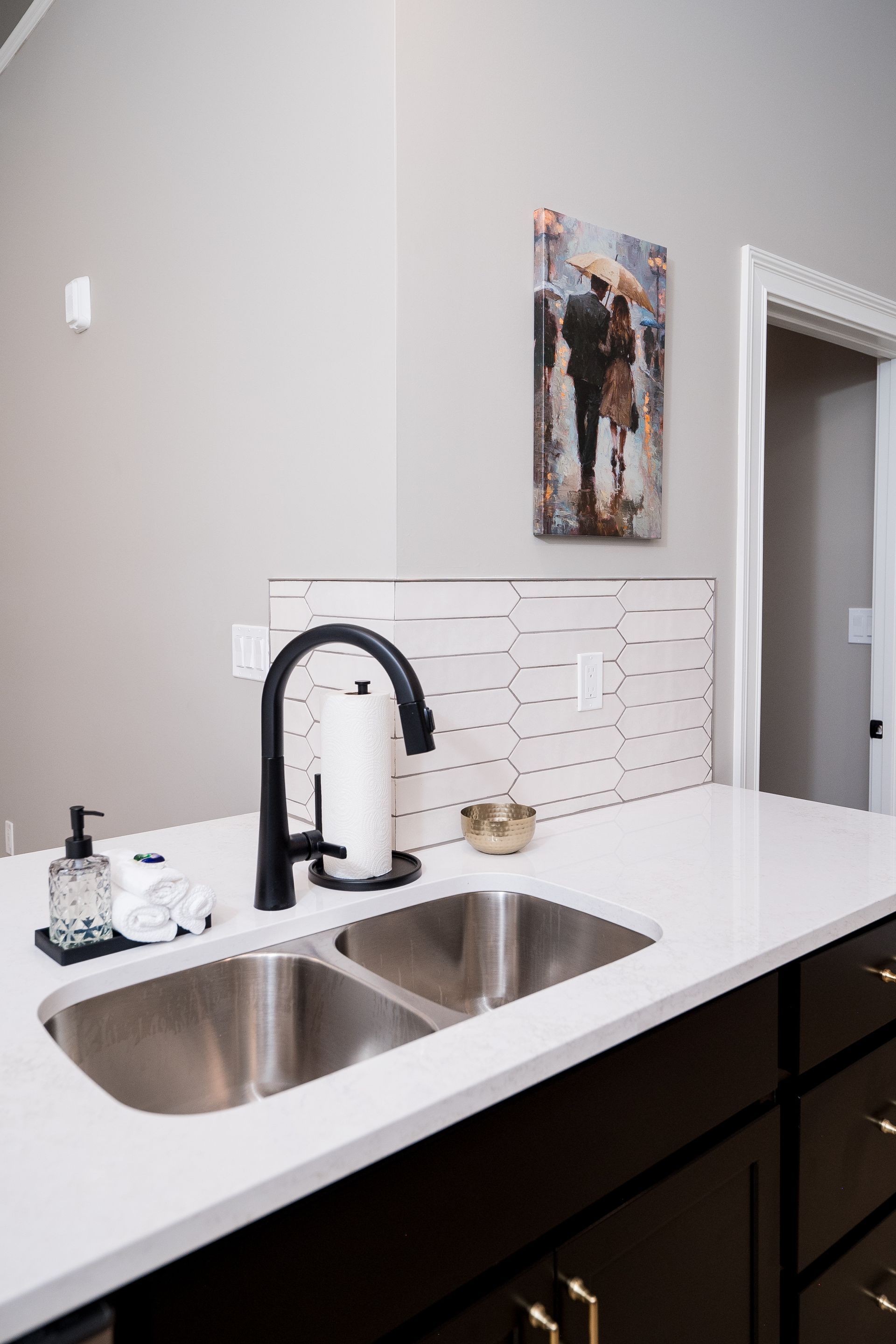 Modern kitchen sink with white countertop, black faucet, dark cabinets, and a small wall painting