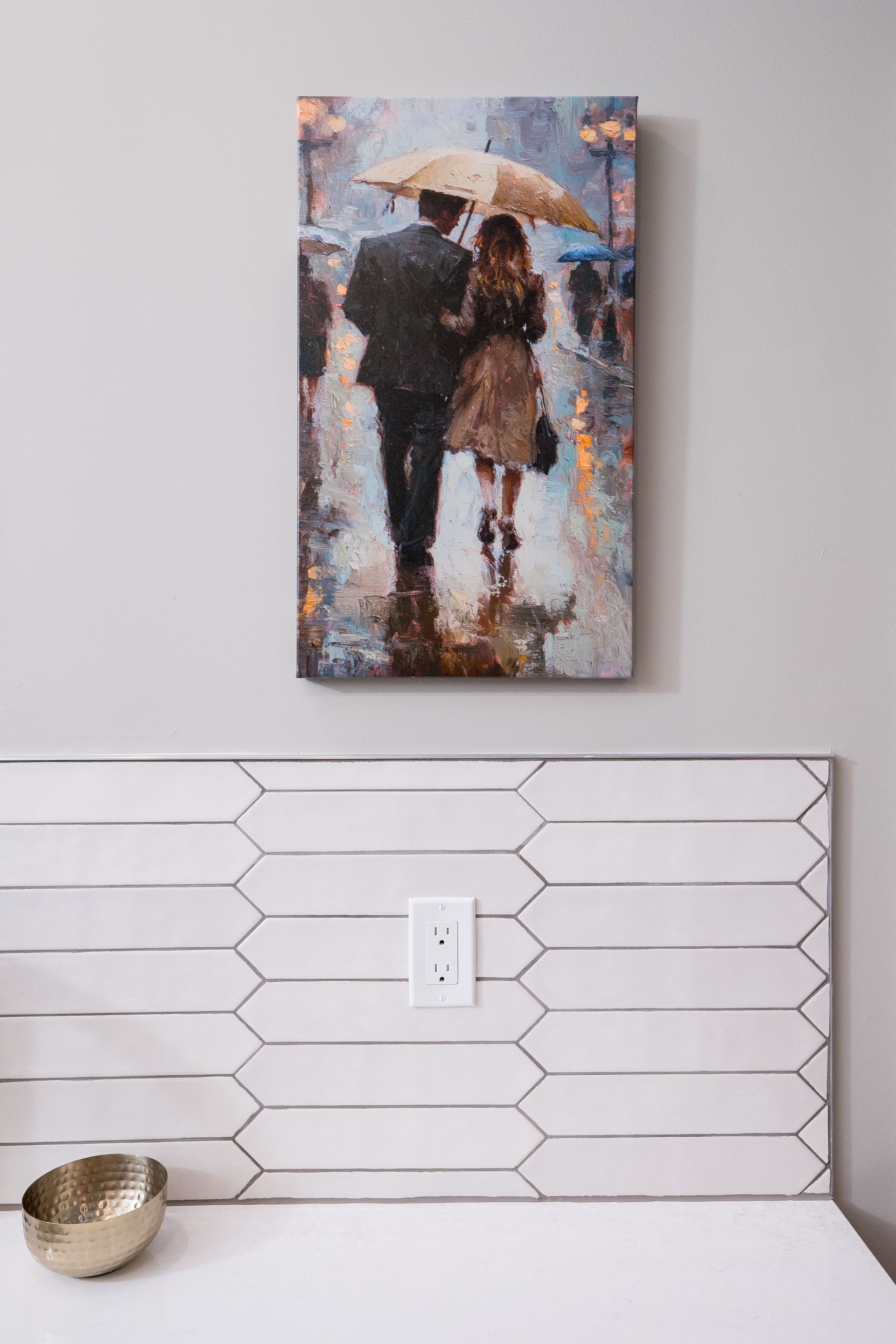 Framed painting of a couple holding an umbrella above a white tiled counter with an outlet and small bowl