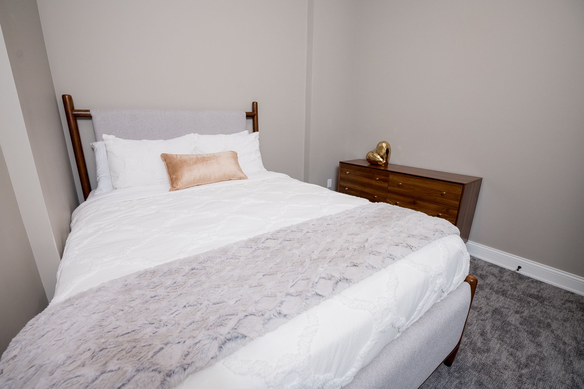 A neatly made white bed in a simple bedroom with a wooden nightstand and gray carpet.