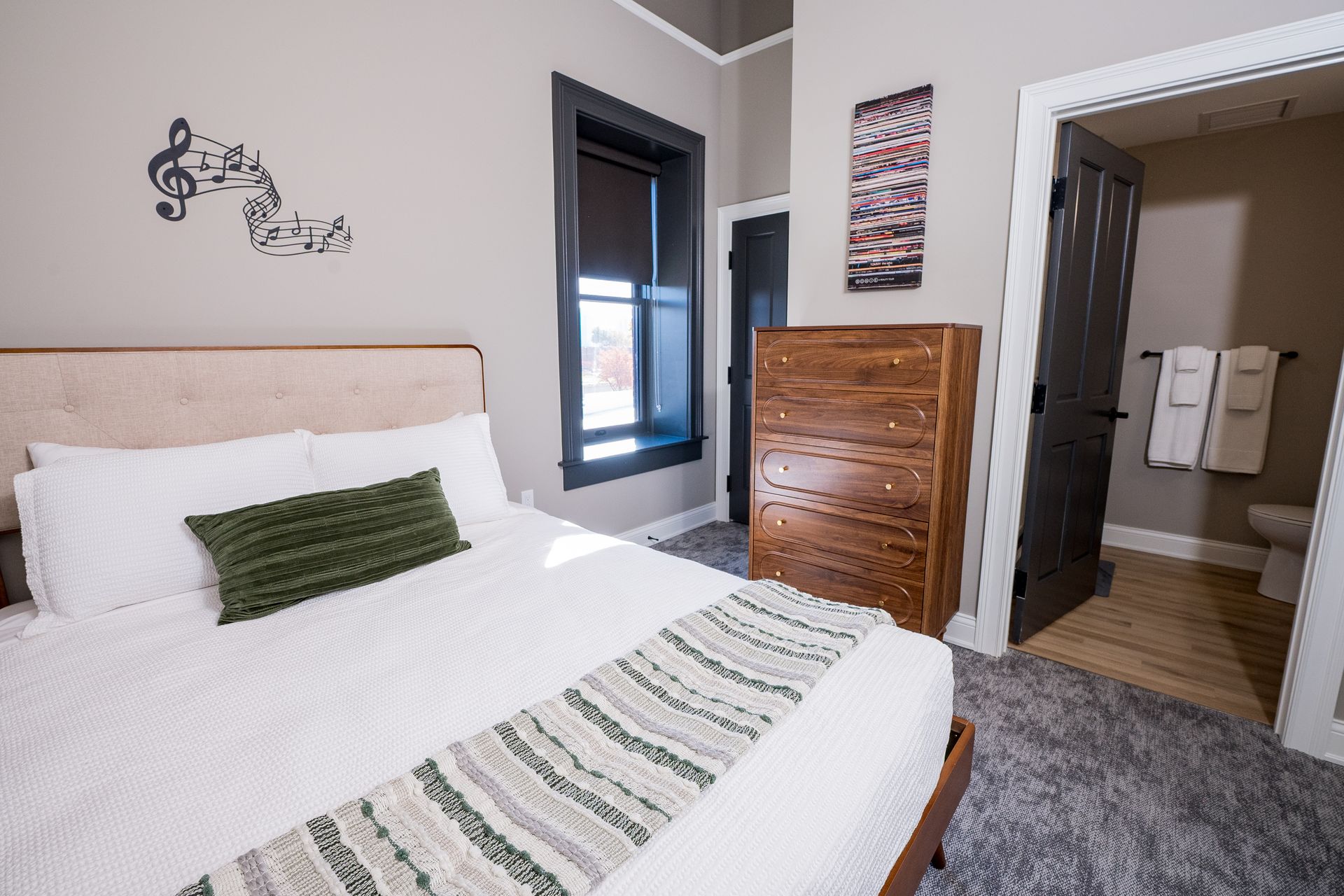Bedroom with white bedding, green accent pillow, wooden dresser, and open bathroom door
