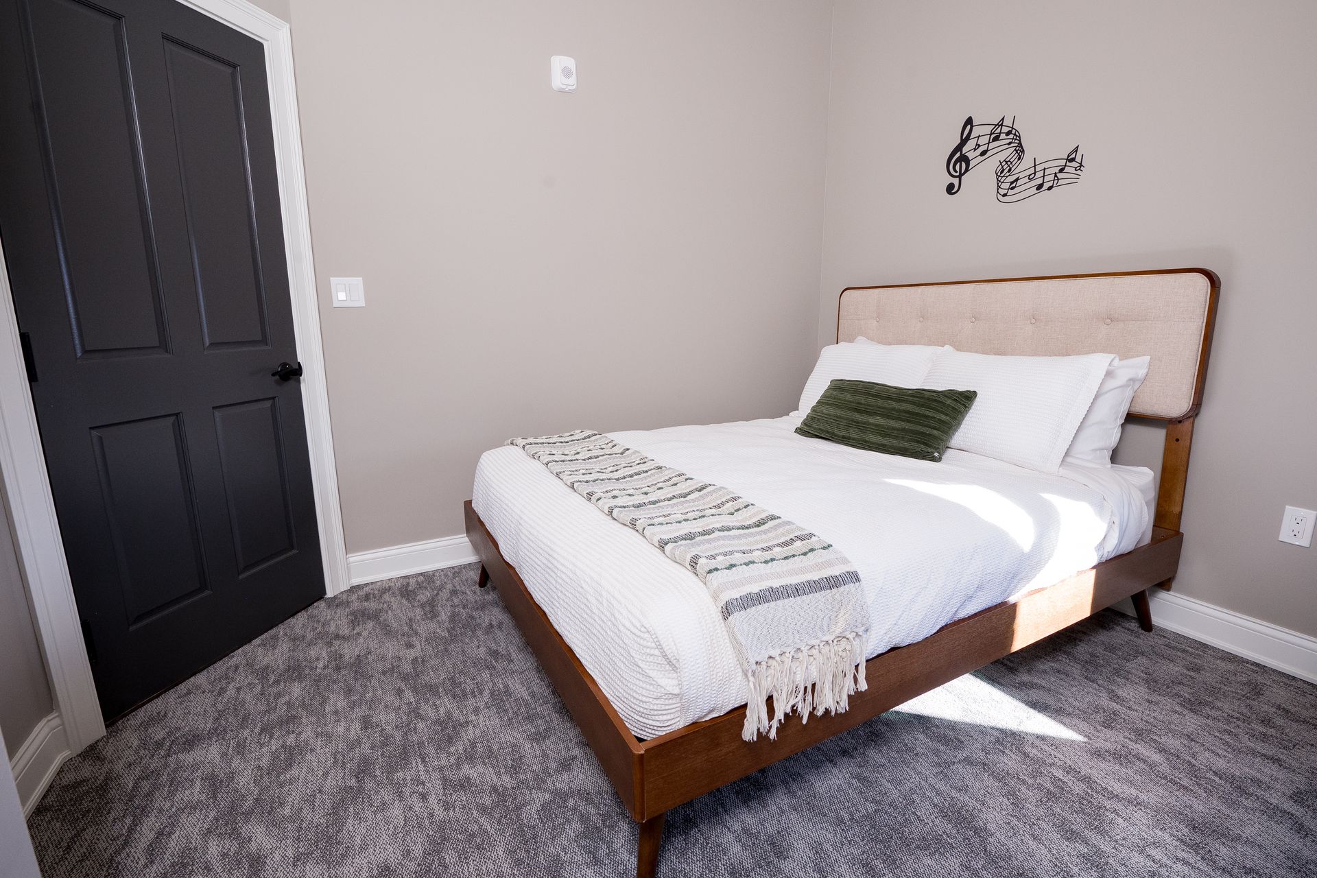 Minimal bedroom with a wooden bed, gray carpet, black door, and light gray walls.