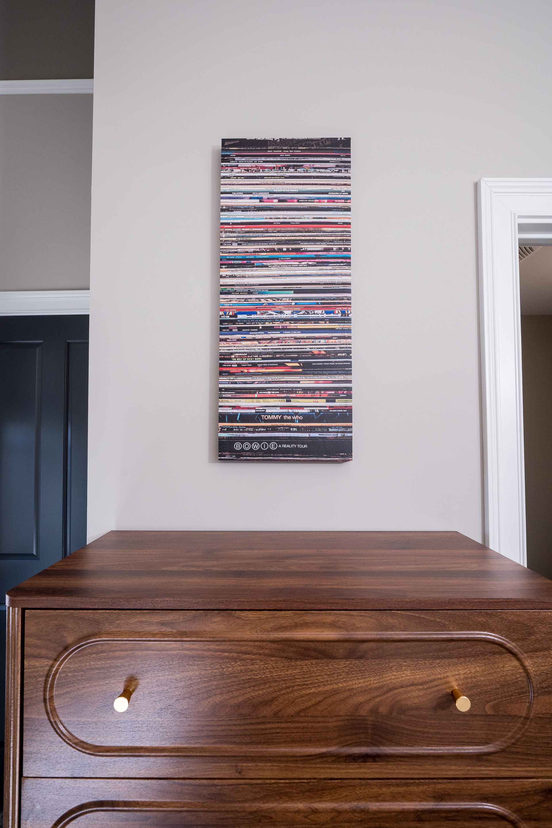 Wooden dresser beneath a narrow abstract wall hanging in a simple room.