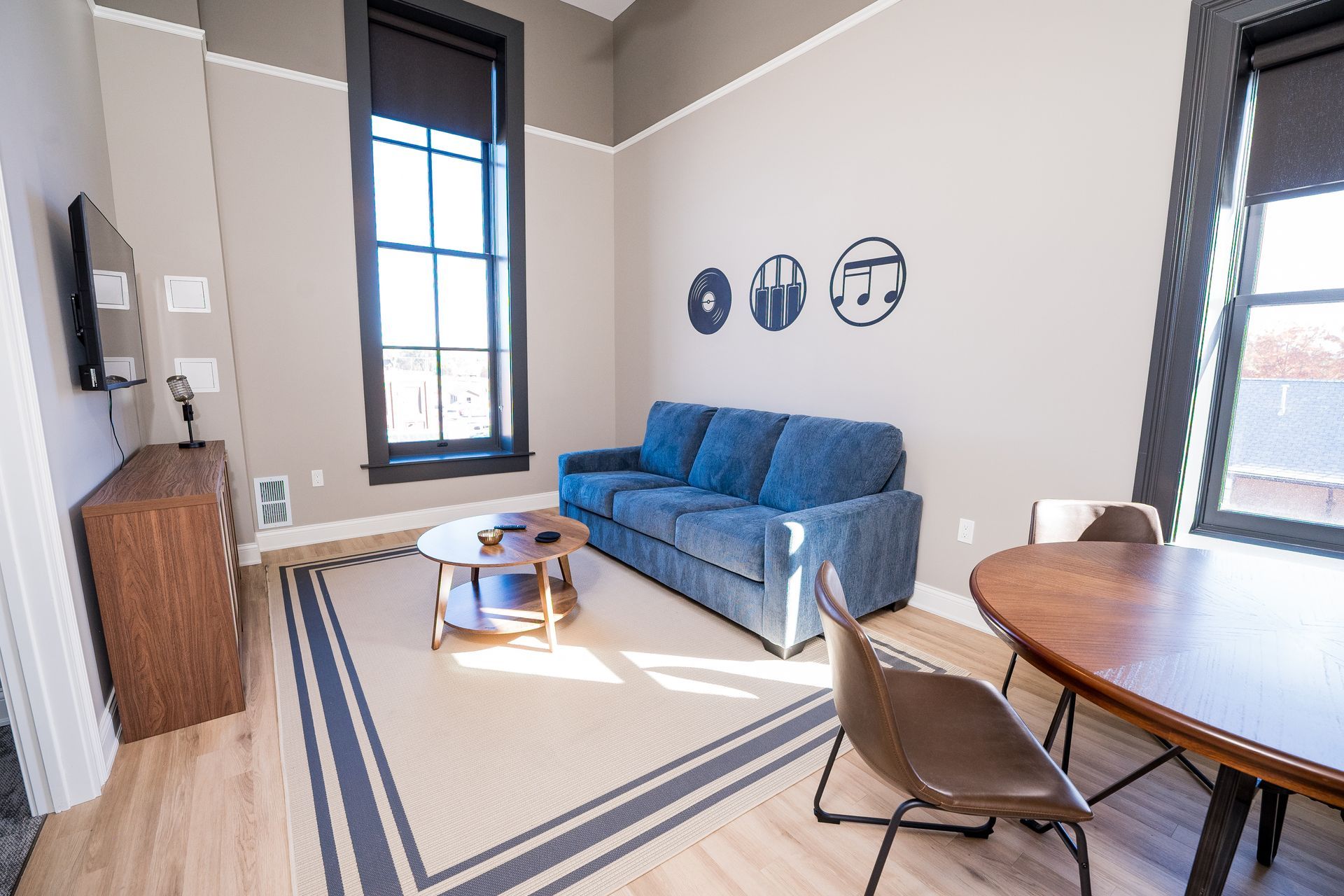 Bright living room with a blue sofa, round coffee table, and dining table by tall windows.