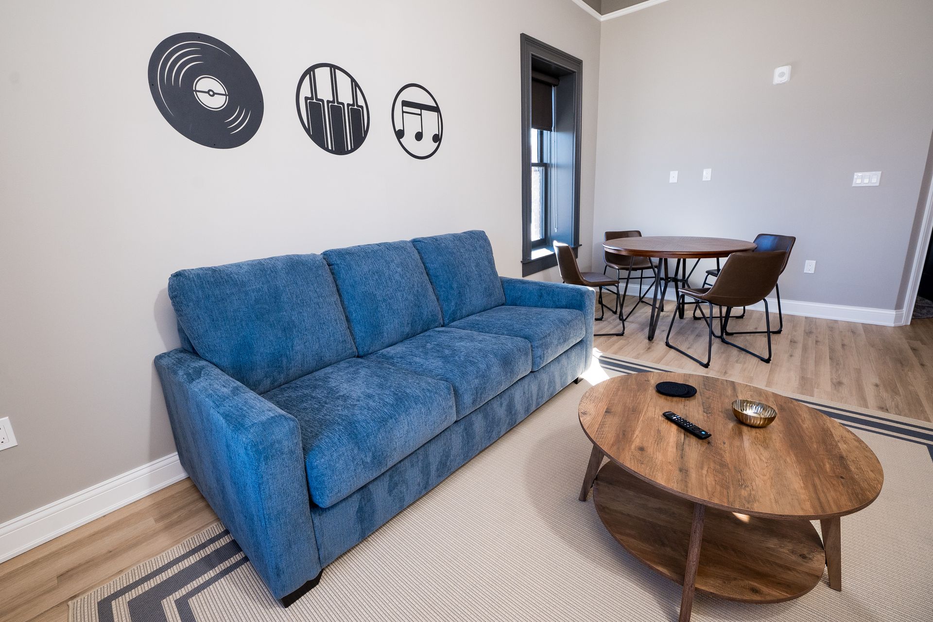 Modern living room with blue sofa, round wooden coffee table, wall art, and dining area in the background