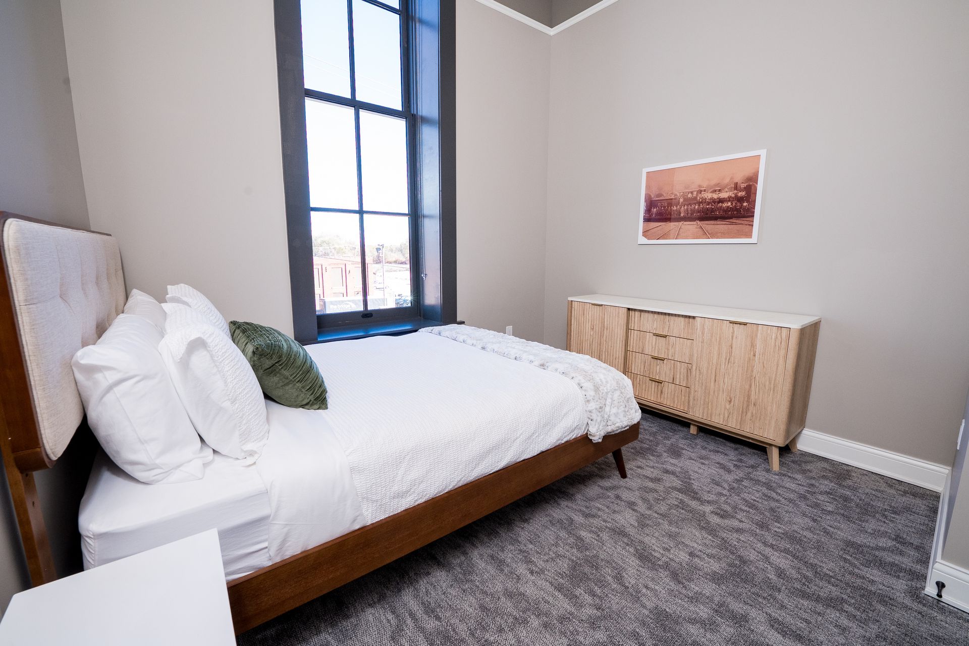 Bright bedroom with white bed, gray carpet, tall window, and light wood dresser beneath wall art