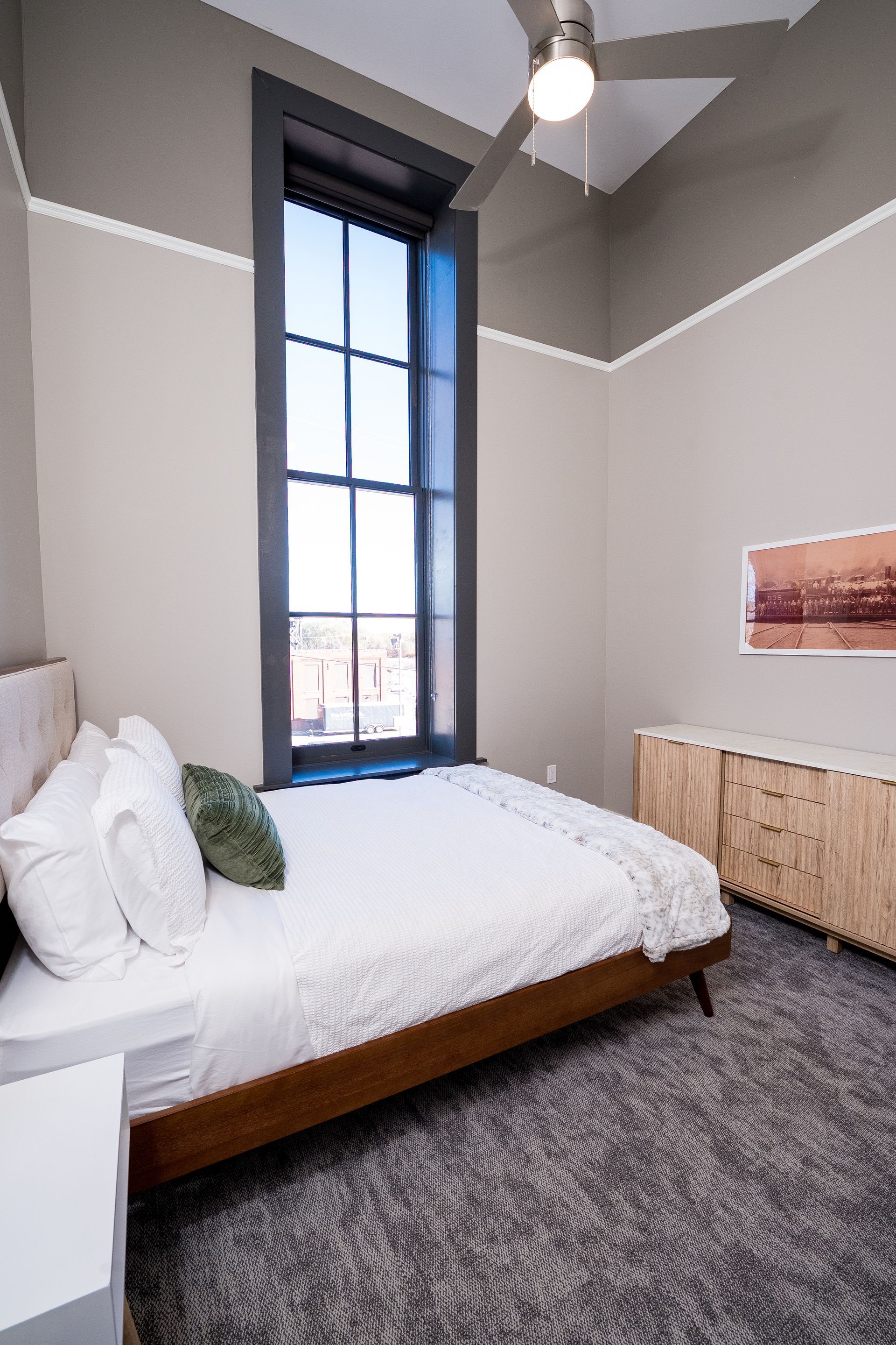 Small bedroom with a white bed, tall window, gray walls, and wood dresser.