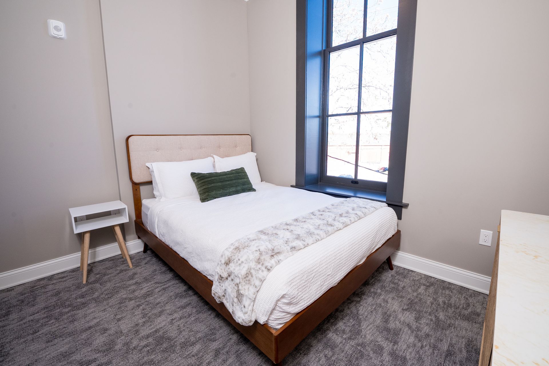 Small bedroom with a bed, side table, gray rug, and tall window.