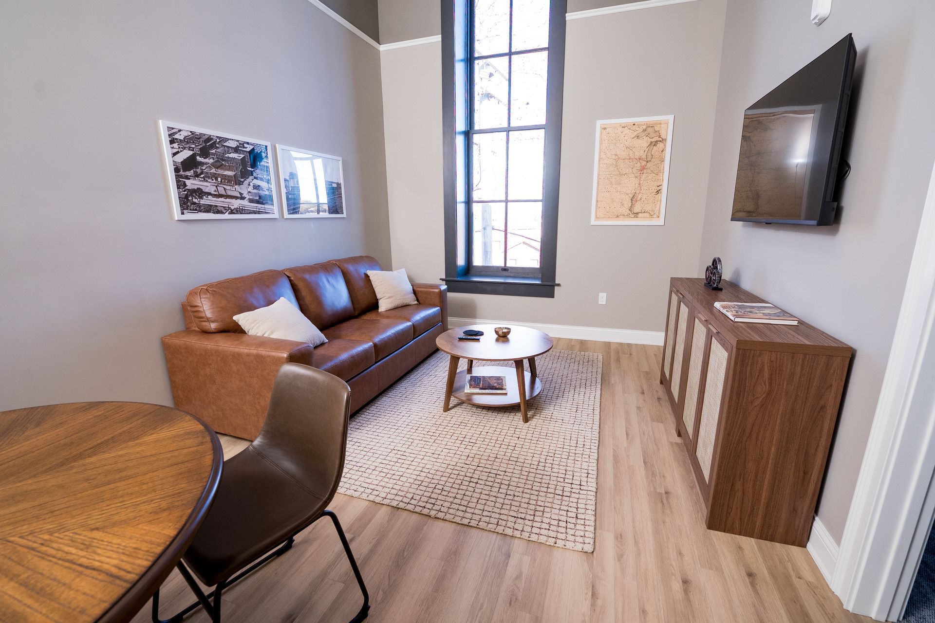 Modern living room with brown sofa, round coffee table, rug, wall art, and large window.