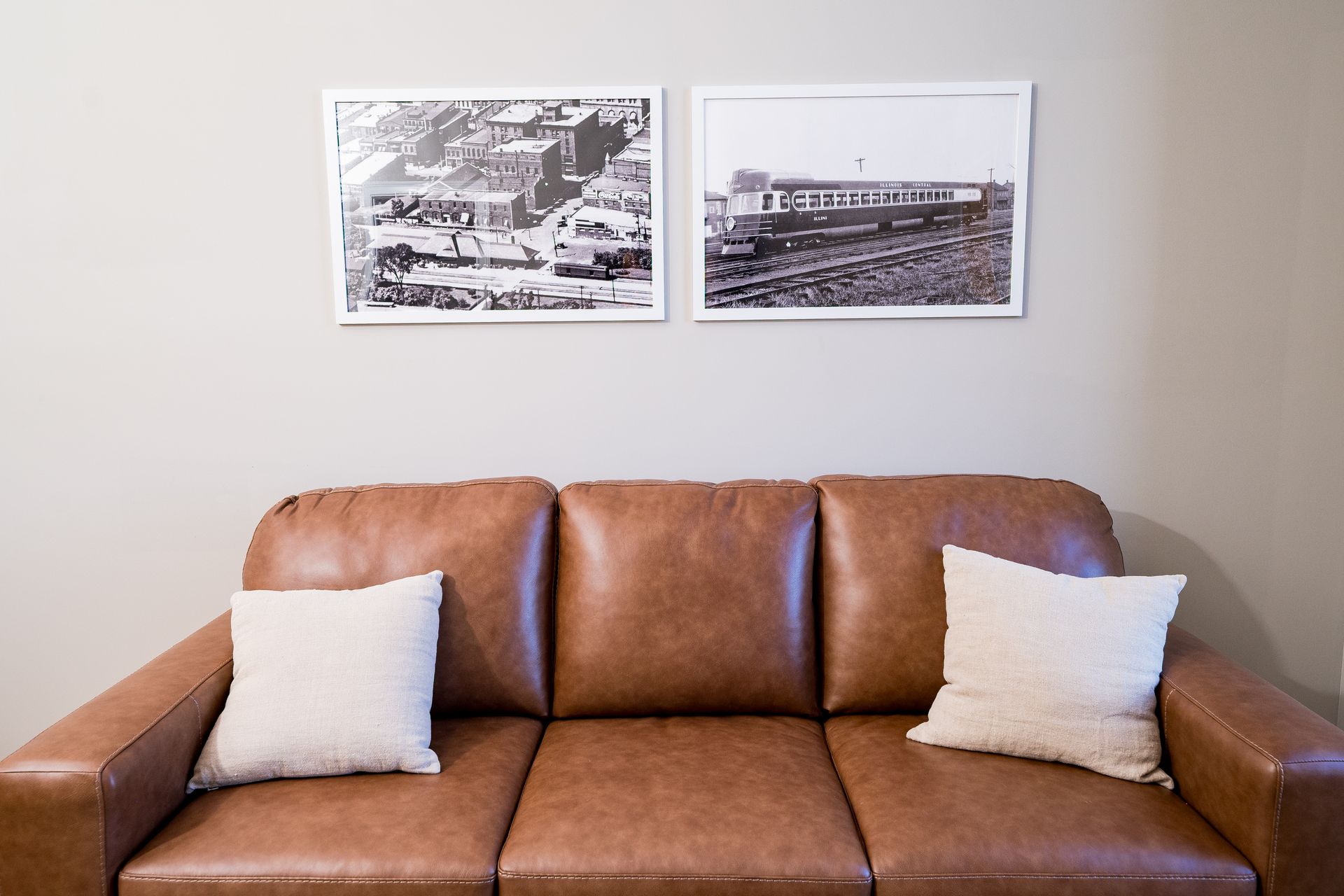 Brown leather sofa with two white pillows beneath two framed black-and-white wall prints