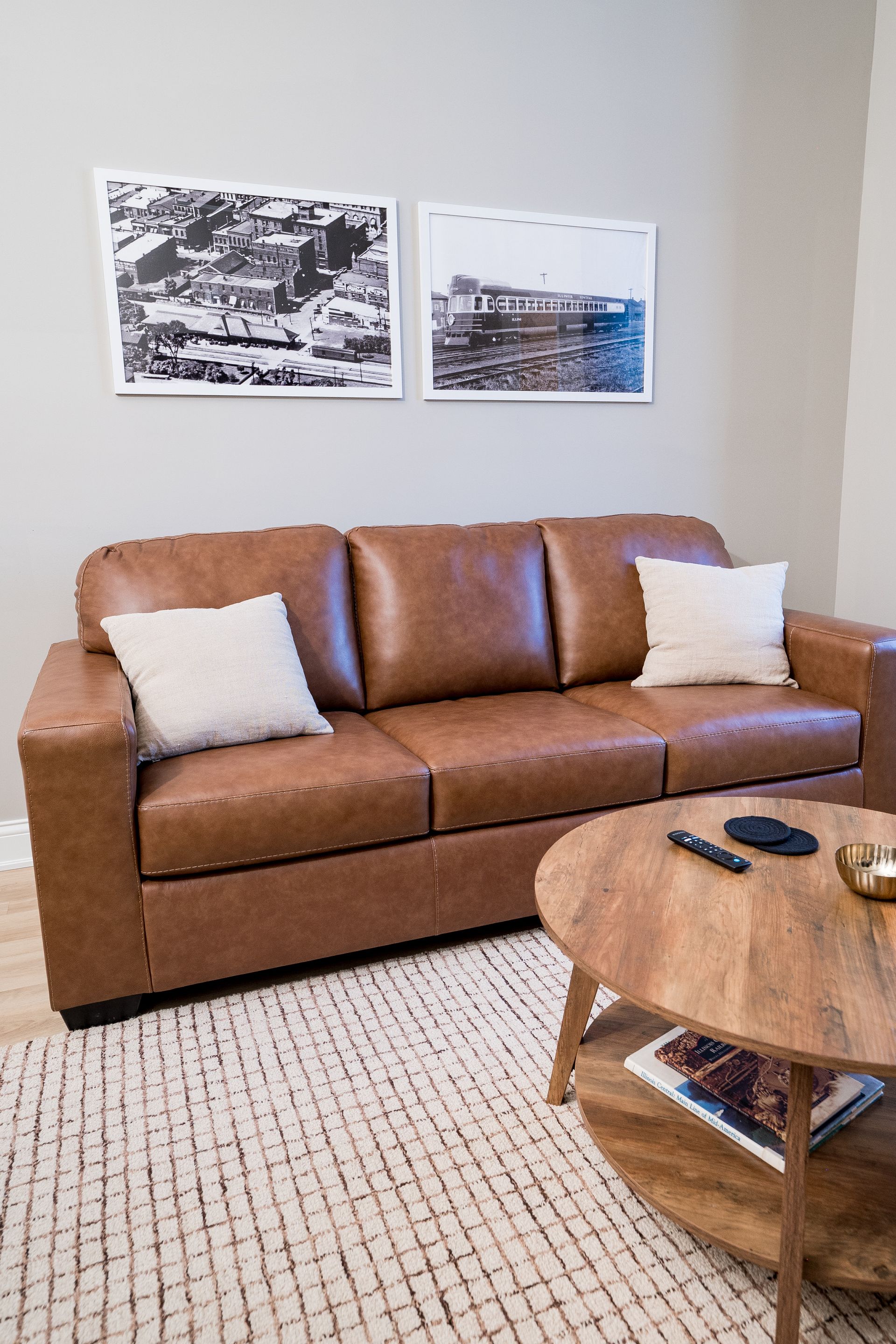 Cozy living room with tan leather sofa, white pillows, framed wall art, and a round wooden coffee table
