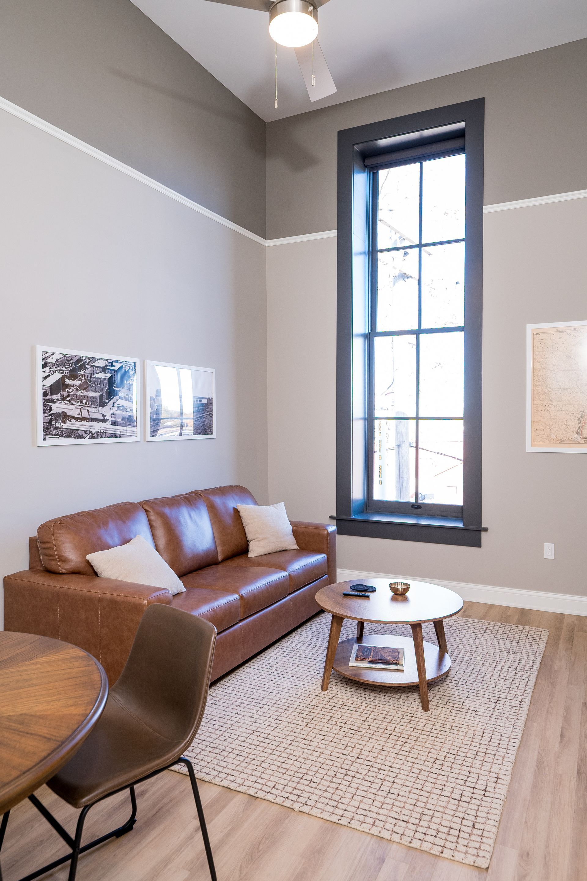 Small living room with a brown leather sofa, round white table, chair, and tall window.