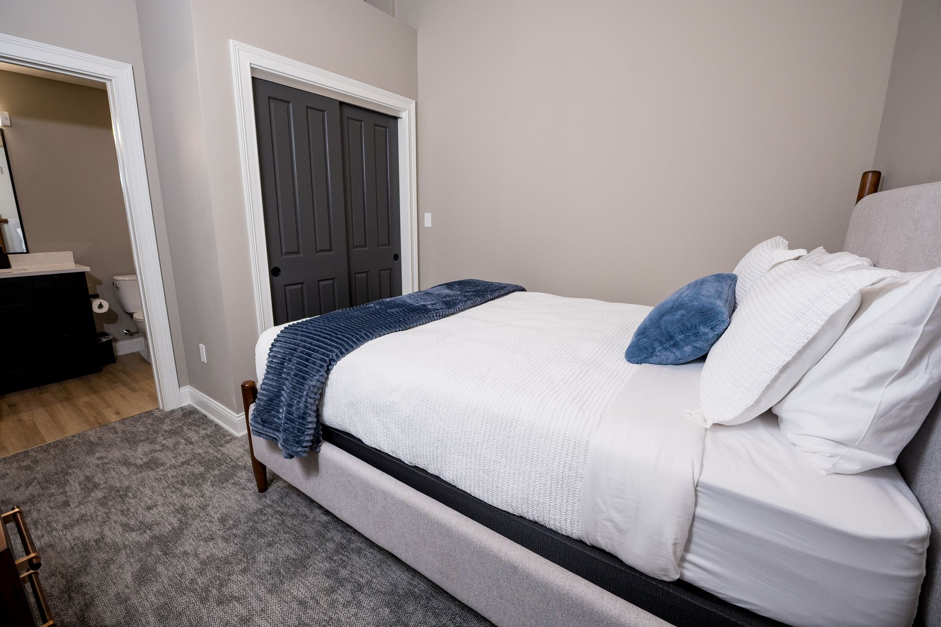 Bright bedroom with a made white bed, blue pillows, gray rug, and open doorway to a bathroom.