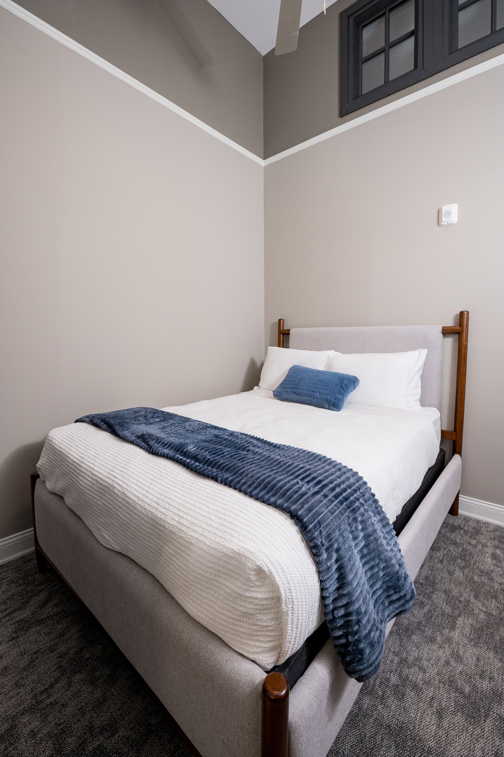 Small bedroom with a neatly made bed, white bedding, blue throw, and a high window.
