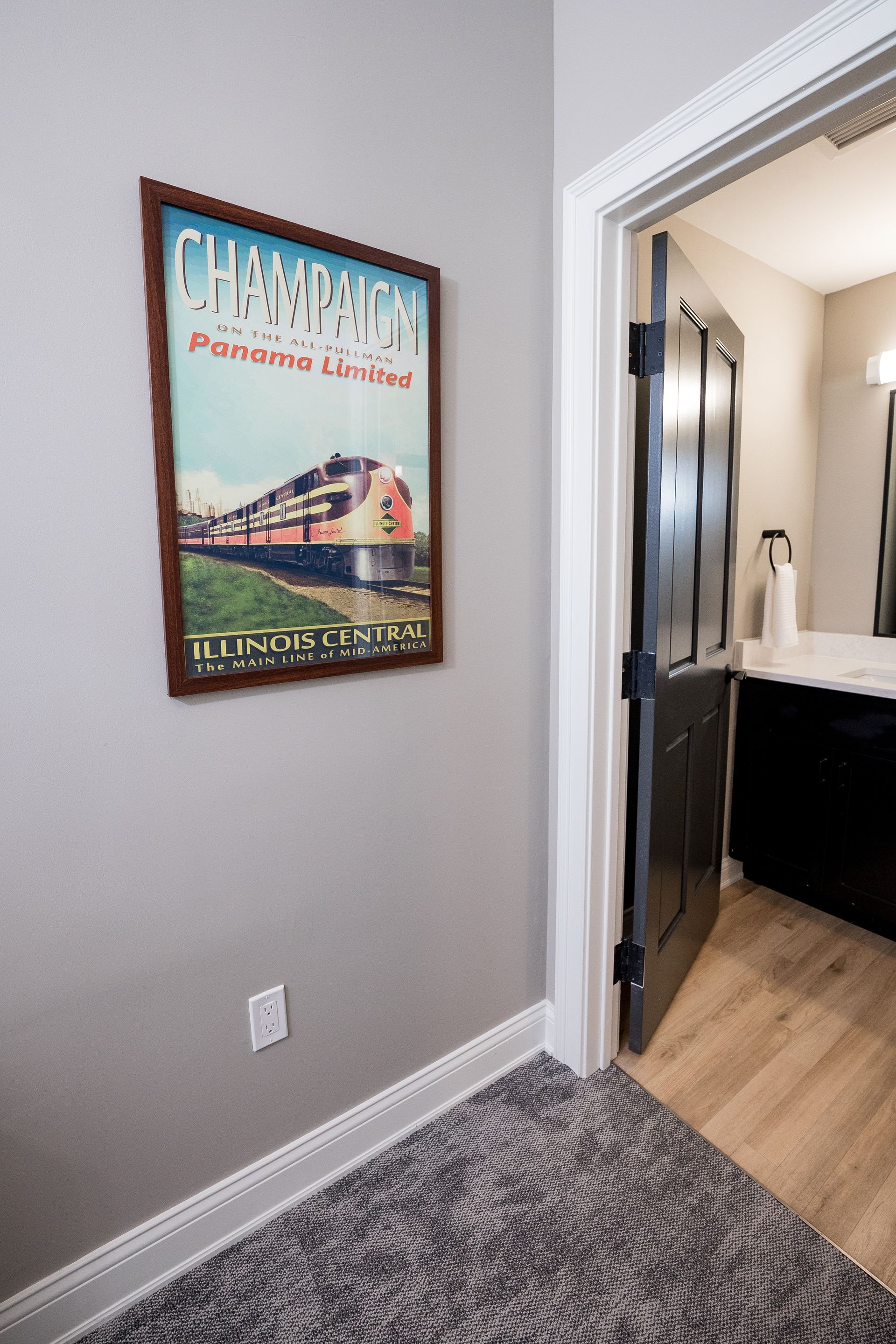 Hallway with framed wall art beside a doorway to a bathroom, gray walls, and wood flooring