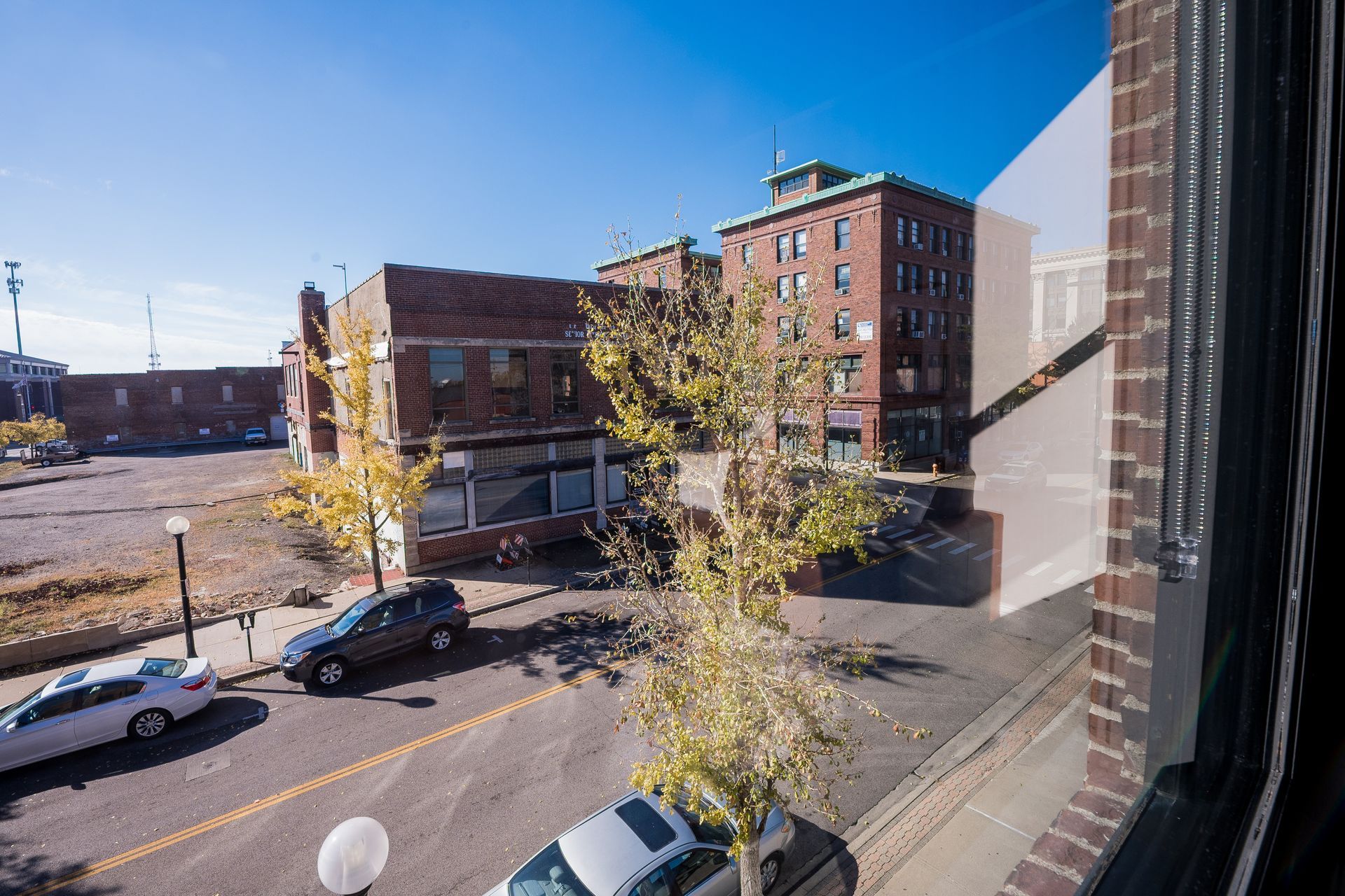Sunny street view from a window, with parked cars, autumn trees, and brick buildings under a blue sky