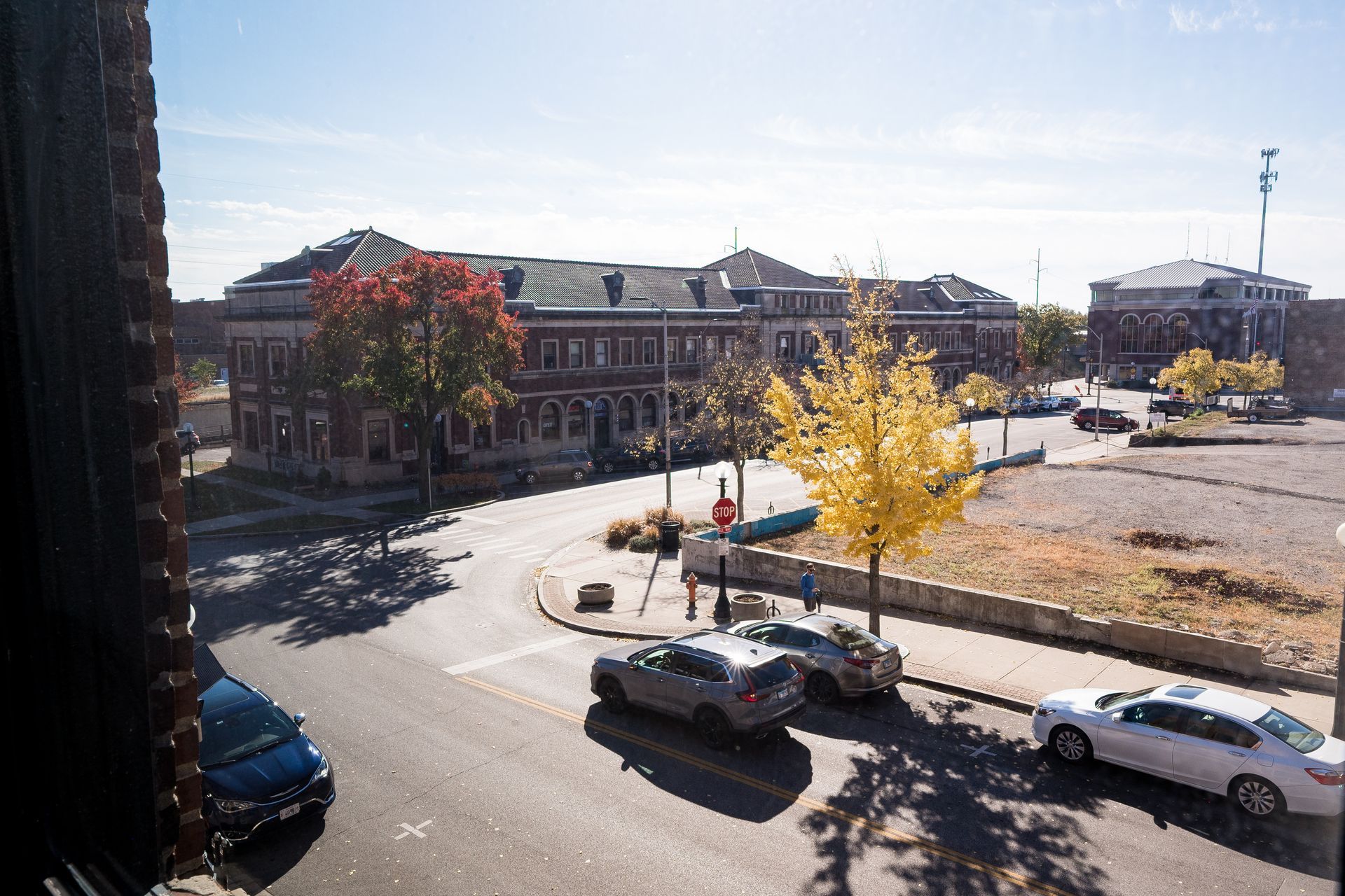 Sunny street corner with parked cars, a brick building, and autumn trees casting shadows.