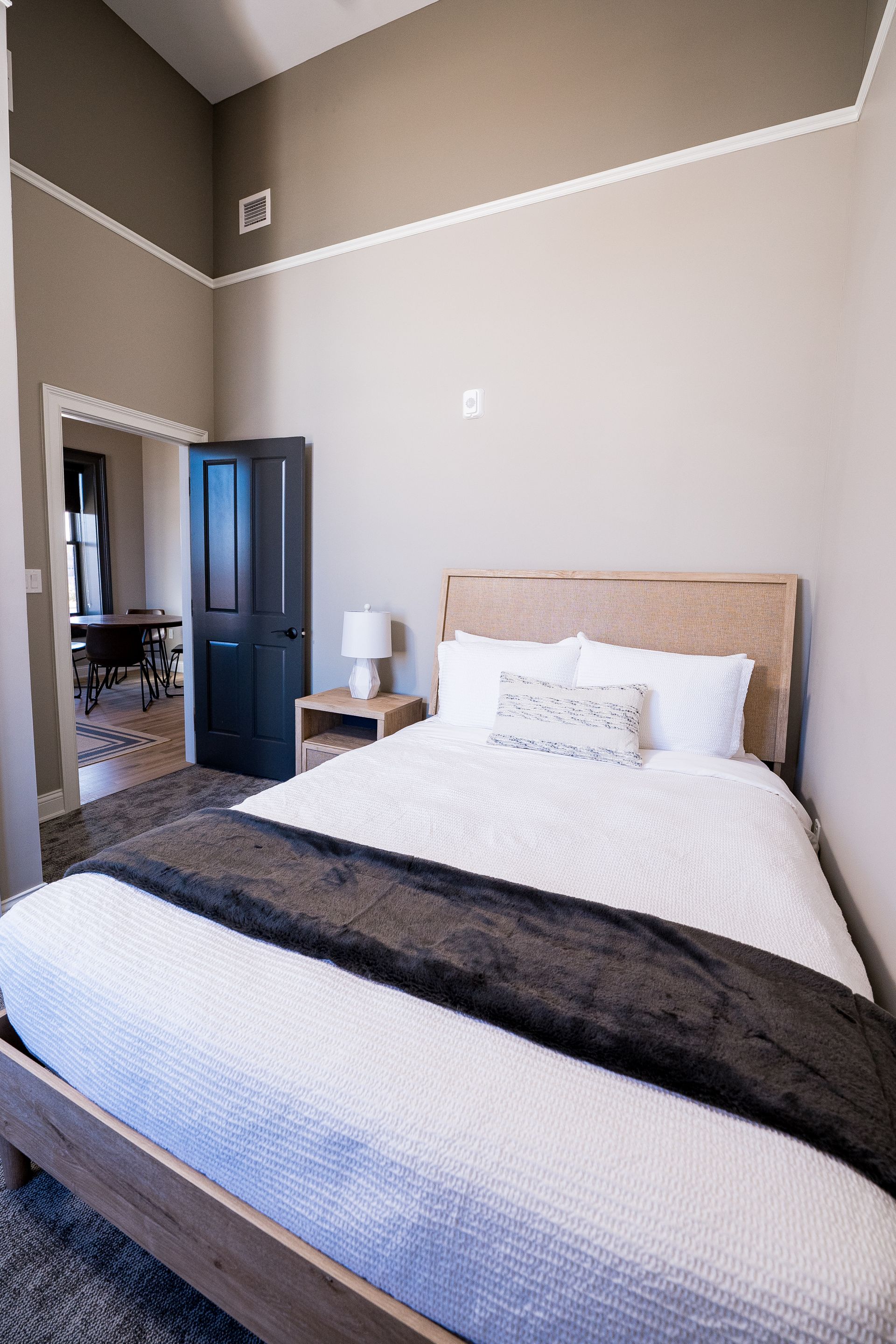 Bright bedroom with a made bed, white bedding, dark throw, and a nightstand by an open door.