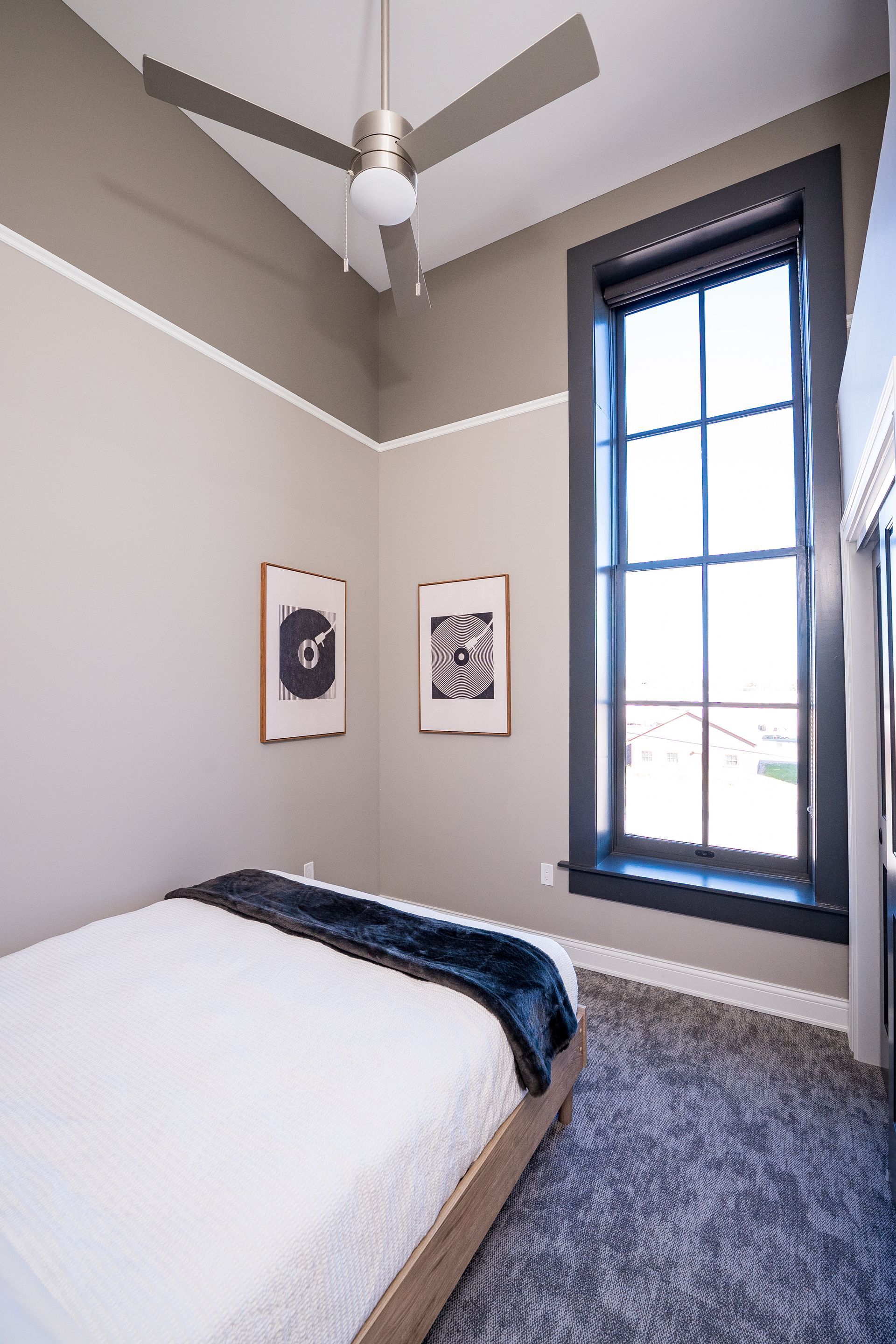 Small bedroom with a bed, ceiling fan, framed art, and tall window.