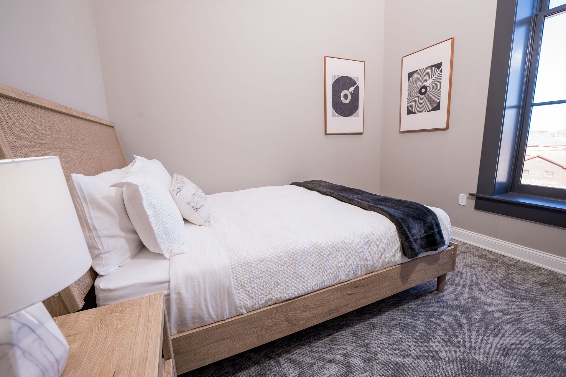 Modern bedroom with a white bed, dark throw blanket, gray rug, and framed wall art by a window.