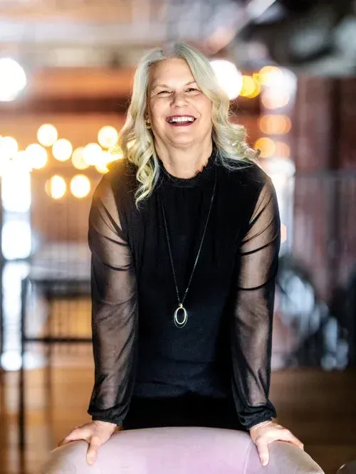 Smiling woman in black outfit seated indoors with warm blurred lights in the background