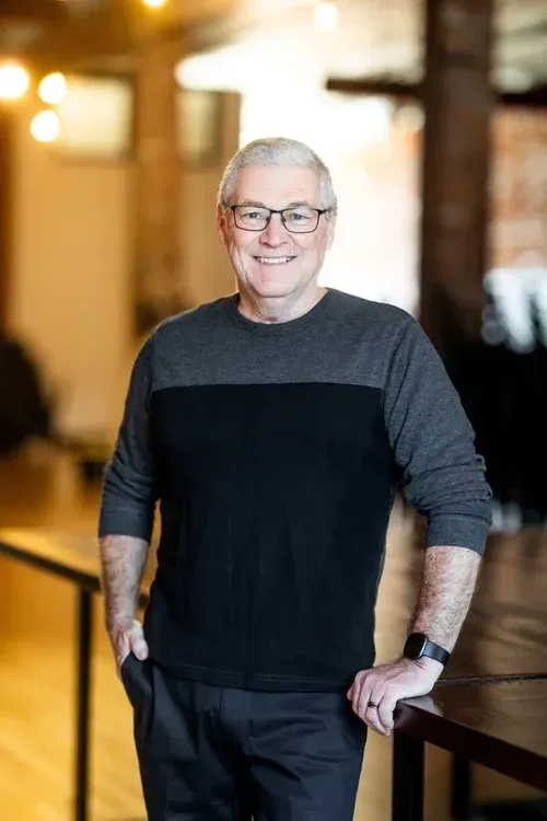 Smiling man in a black and gray sweater leaning on a table in a softly lit indoor hallway