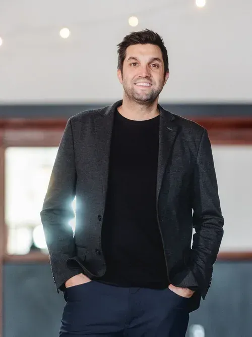 Man standing indoors in a dark blazer and black shirt, hands in pockets, smiling at the camera