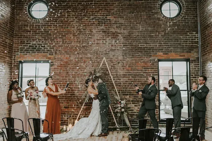 Wedding party and couple exchanging vows in front of a brick wall and wooden triangle backdrop