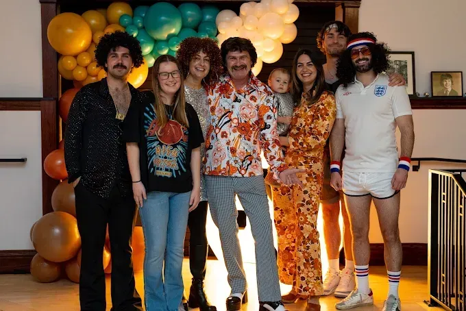 Six people posing indoors in front of a colorful balloon arch, smiling at a party.