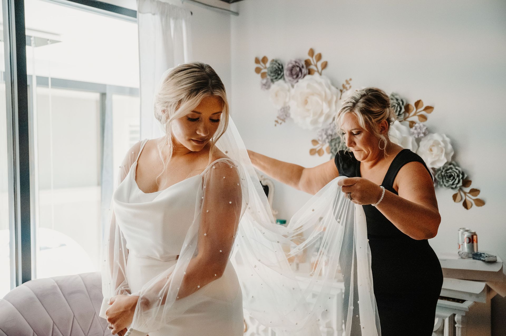 Bride in white dress with veil being helped by attendant in black dress in a bright room