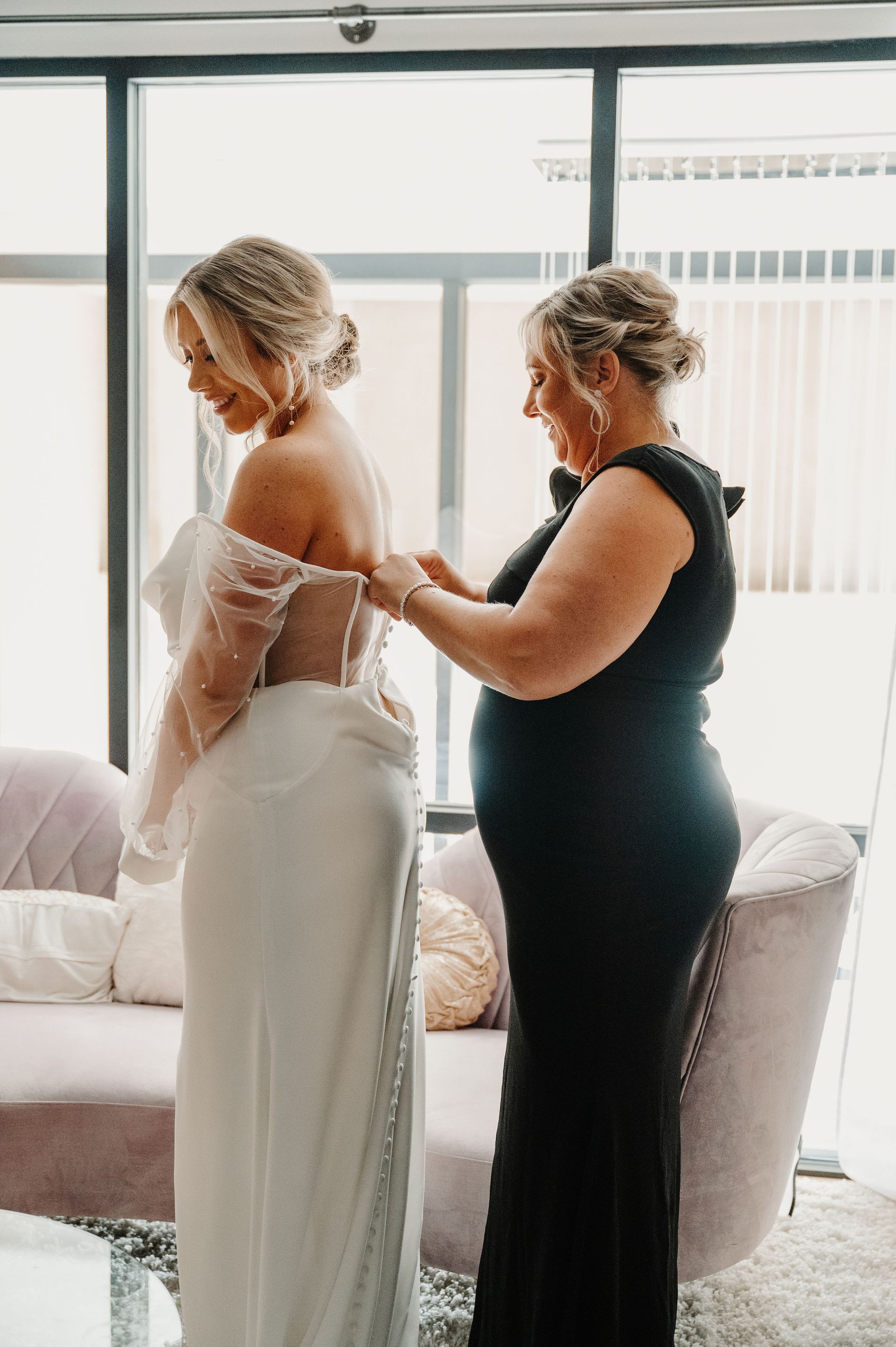 Woman in a white dress being buttoned by another woman in a black dress by a bright window